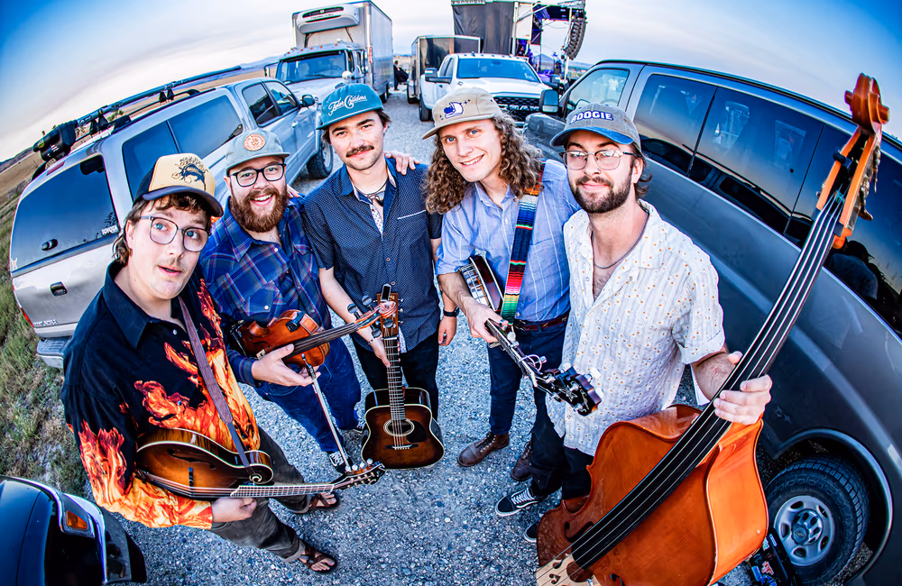 Five smiling musicians holding string instruments, standing on gravel with parked trucks and vans behind them.