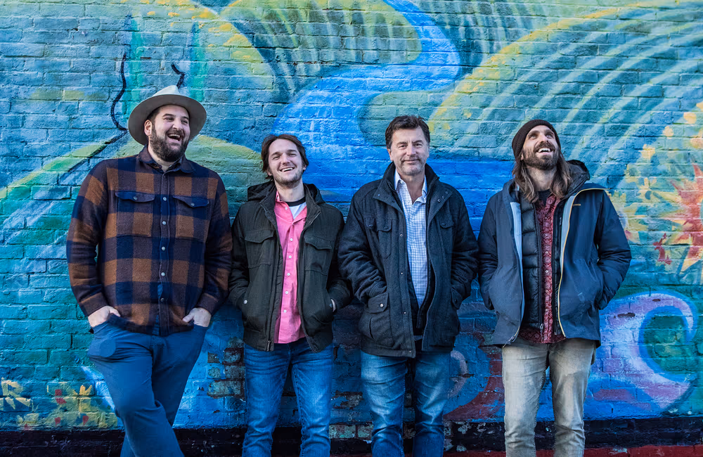 Four men standing and smiling in front of a colorful graffiti-covered brick wall.