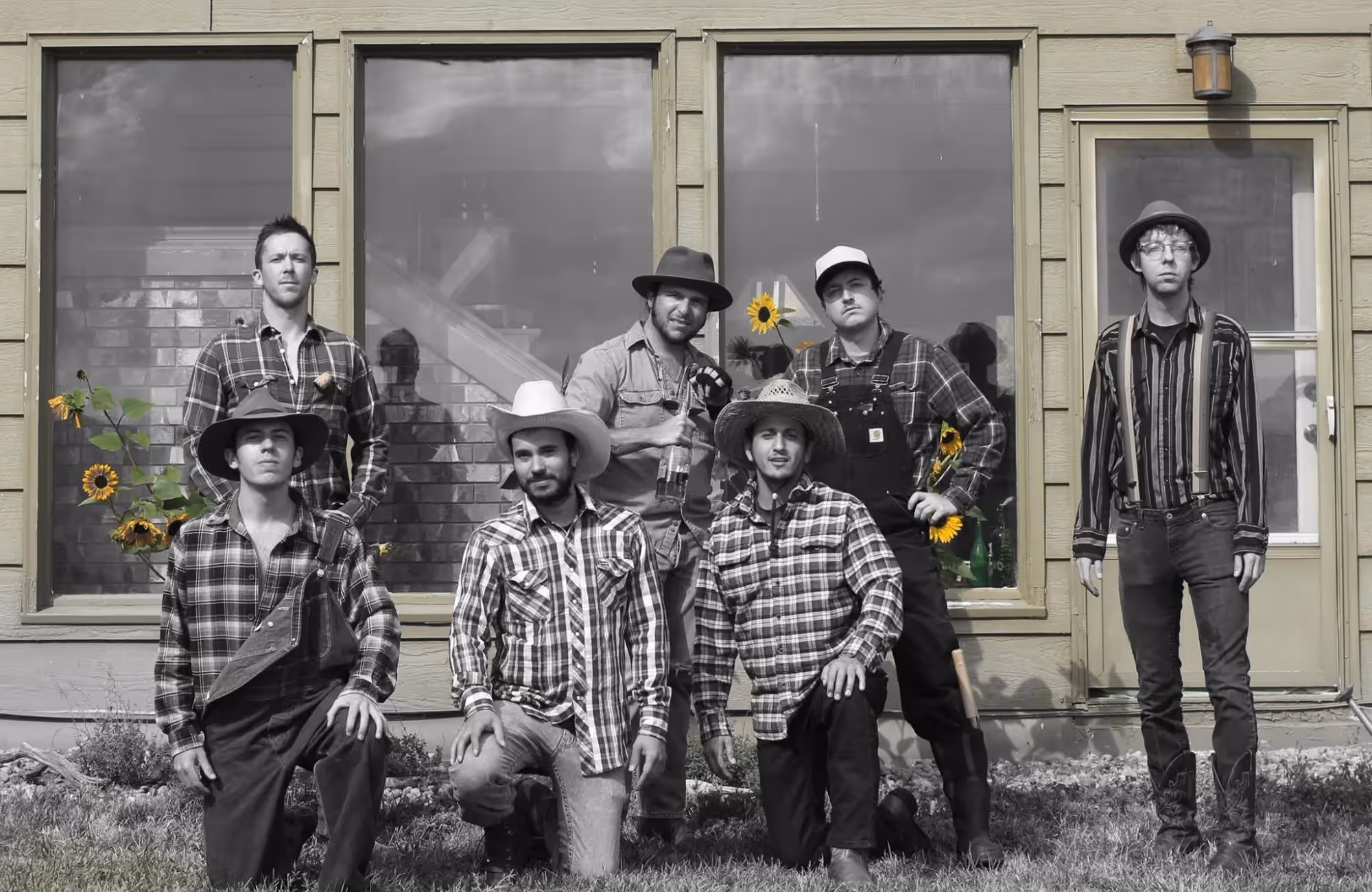 Group of six men in casual farm-style attire, some wearing hats, posing outside a house with sunflowers in the background.