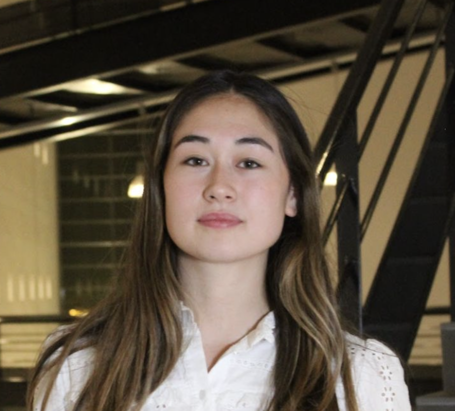 Jeune femme aux cheveux longs et bruns portant une blouse blanche, posant devant un escalier métallique dans un environnement intérieur moderne.