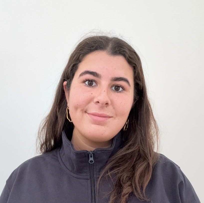 Portrait d'une femme souriante aux cheveux longs bruns, portant une veste à fermeture éclair et des boucles d'oreilles dorées.