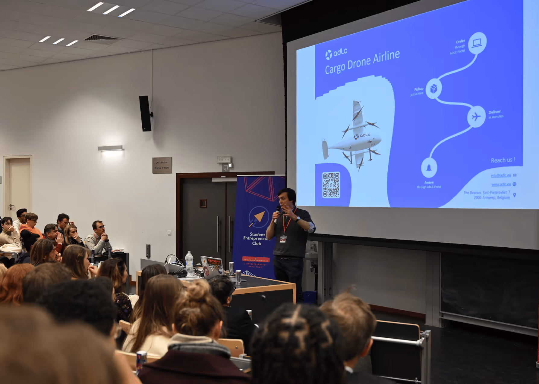Un conférencier présente un projet de drone cargo à une salle pleine d'étudiants attentifs dans un amphithéâtre.