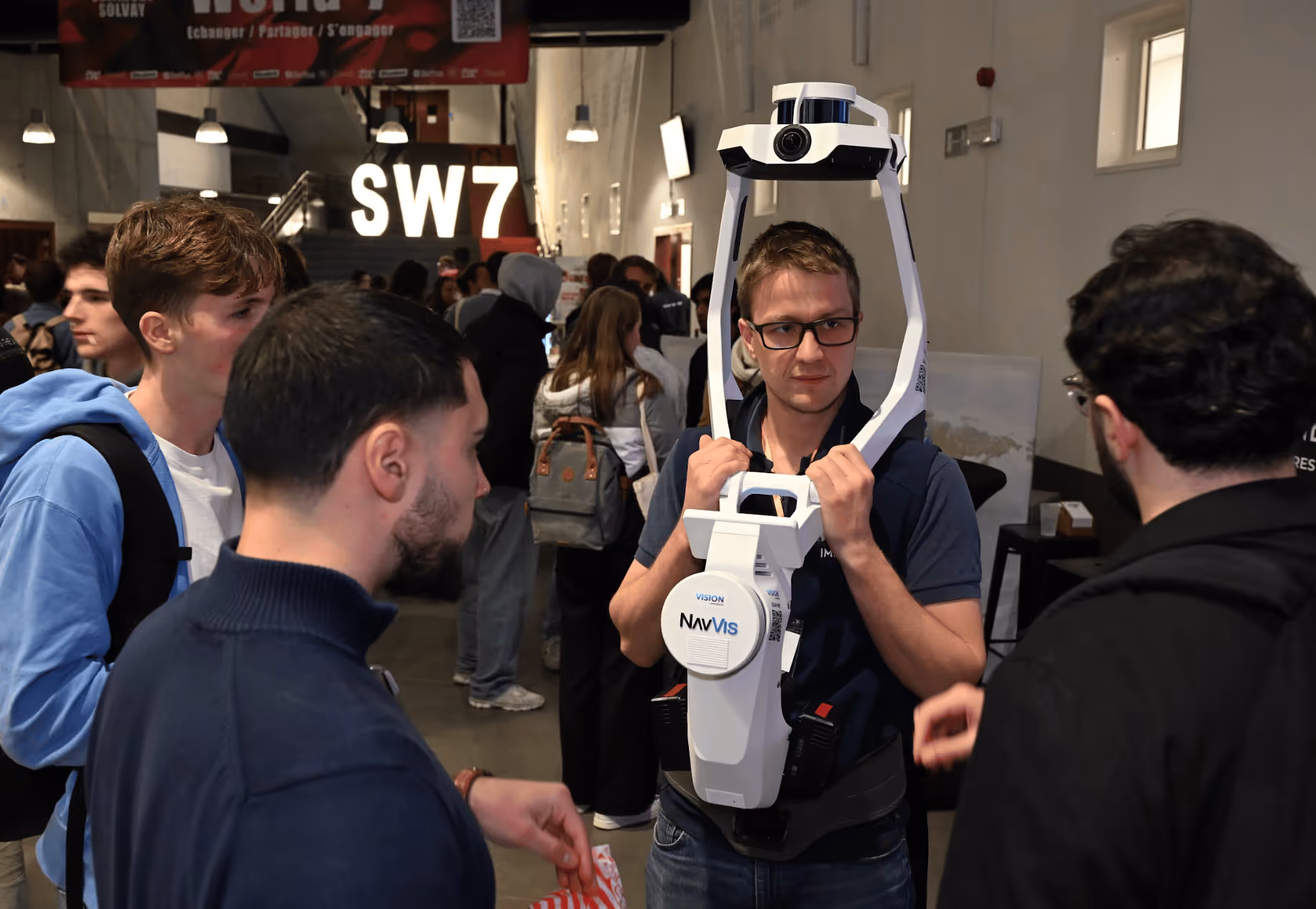 Un homme portant un appareil de capture 3D NavVis discute avec plusieurs personnes lors d’un événement intérieur.