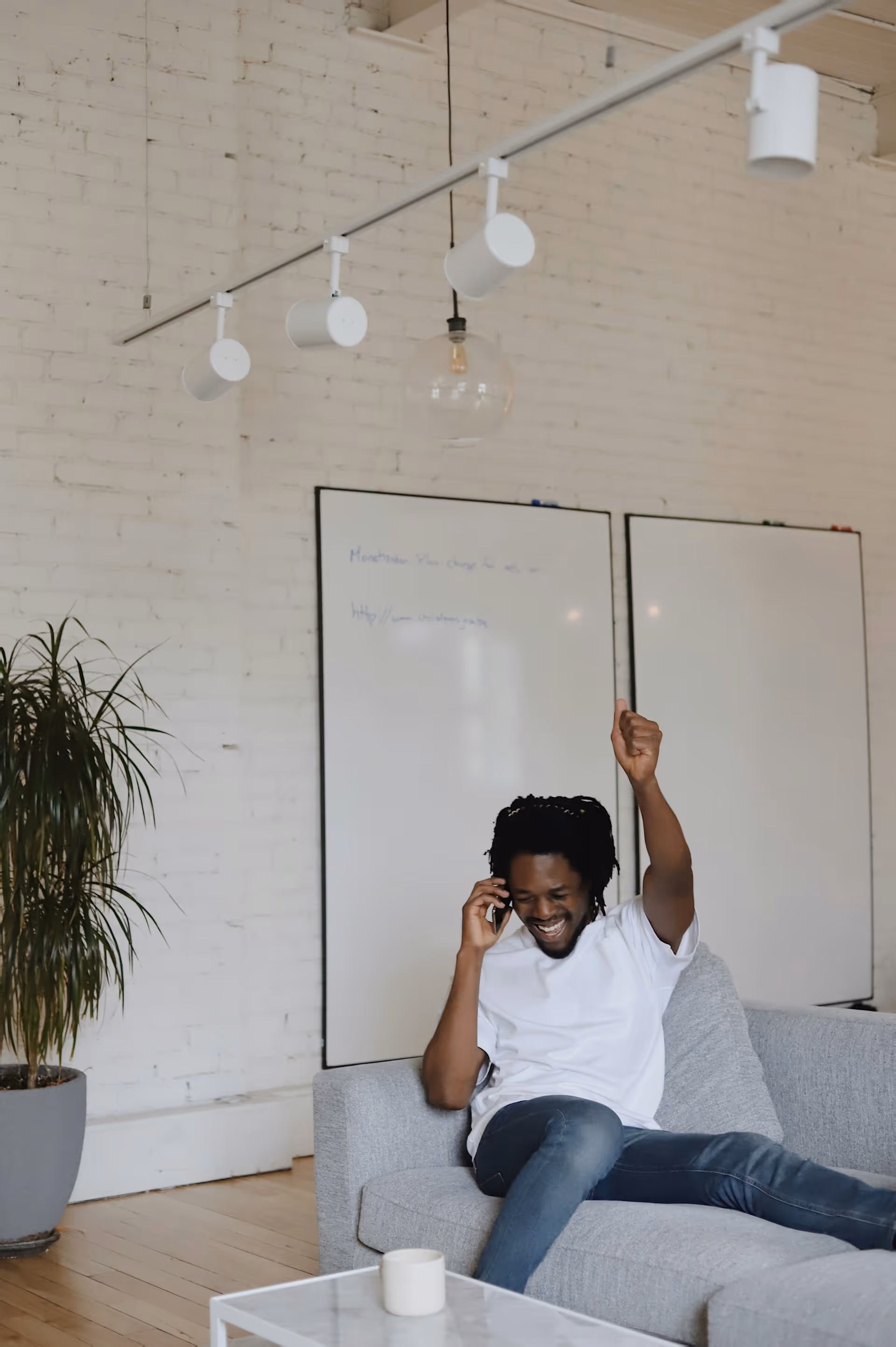 Un homme assis sur un canapé gris, parlant au téléphone avec enthousiasme et levant le pouce en l'air dans un bureau moderne avec tableaux blancs et une plante.