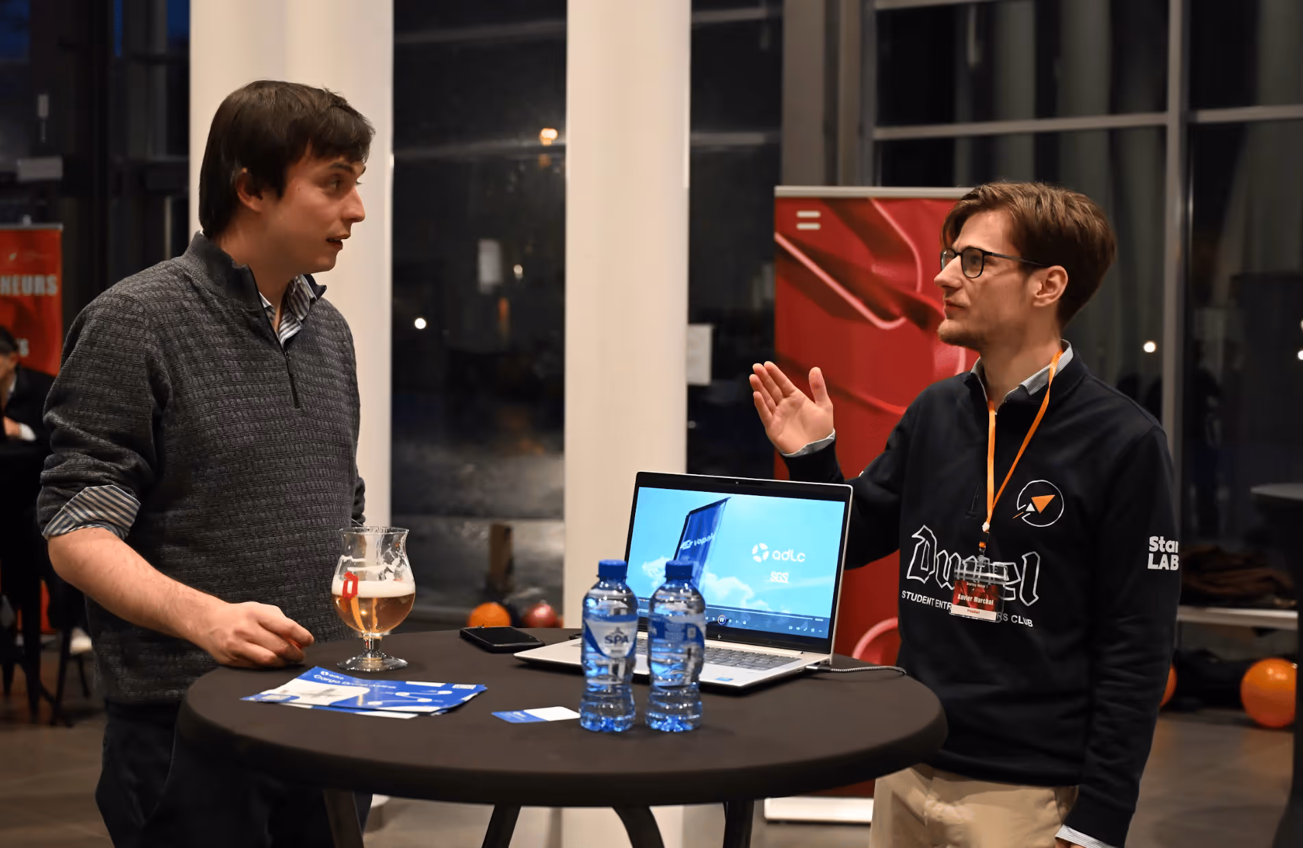 Deux hommes discutent debout à une table haute avec un ordinateur portable, des bouteilles d'eau et une bière dans un cadre intérieur.