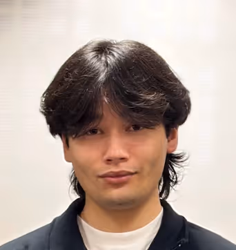 Portrait d'un jeune homme avec des cheveux noirs mi-longs et une expression neutre, portant une veste noire sur un fond clair.