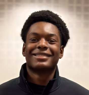 Portrait d'un jeune homme souriant portant un haut noir avec un col montant, sur un fond flou clair.