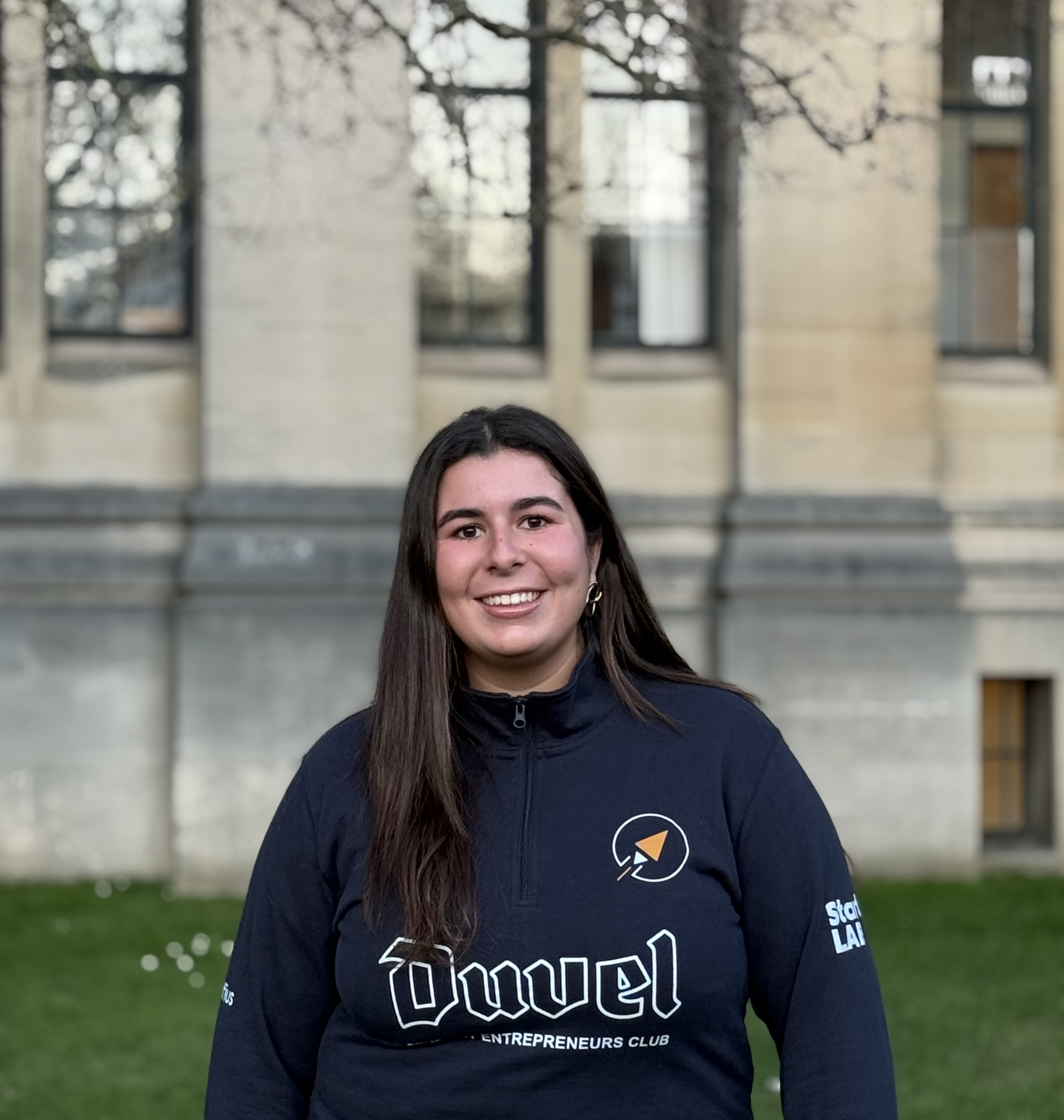 Une femme souriante portant un sweat-shirt bleu marine avec le logo et le texte 'Ouwel Entrepreneurs Club' debout devant un bâtiment en pierre.