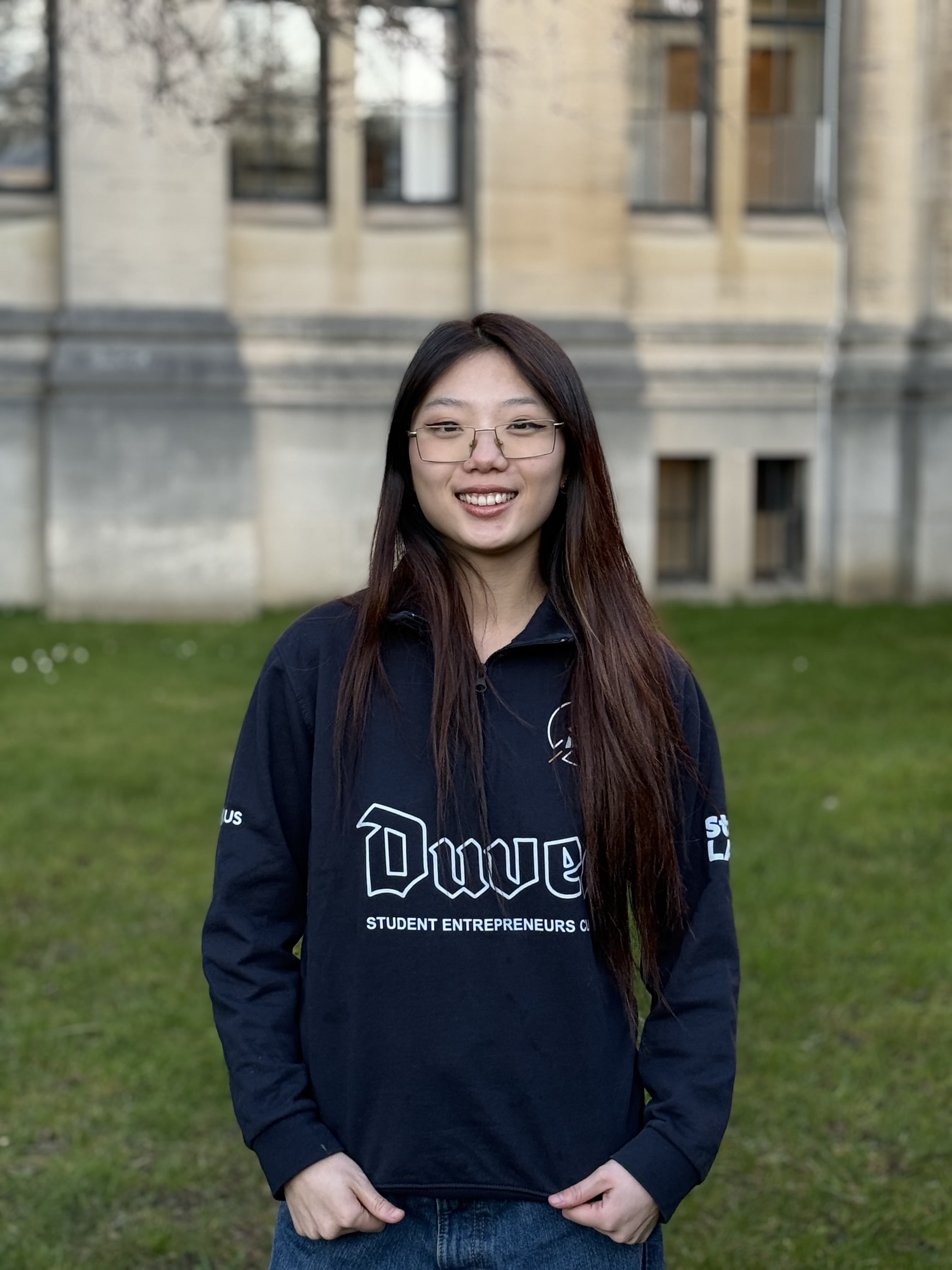 Femme asiatique souriante portant des lunettes et un haut noir, devant un mur clair flou.