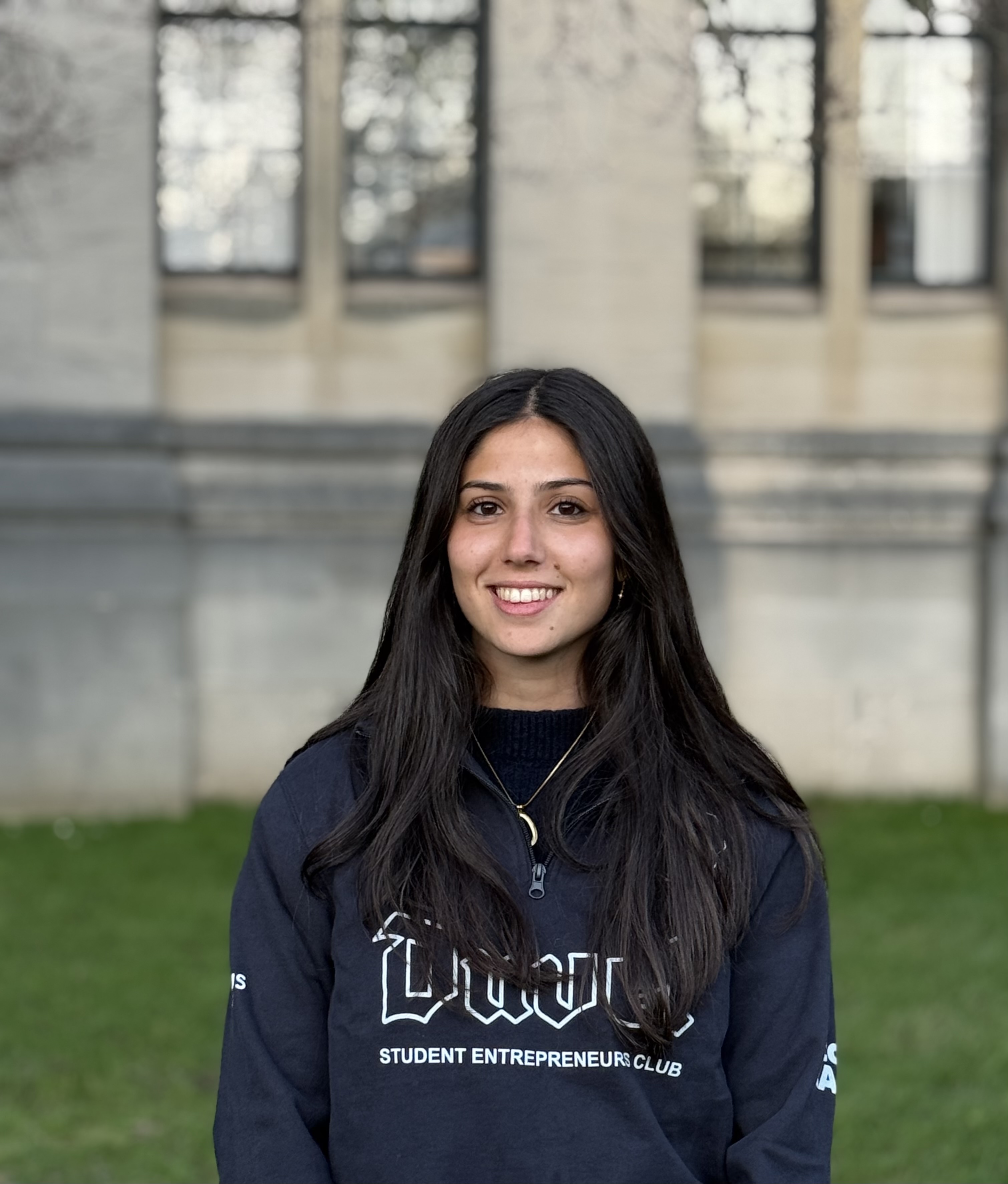 Jeune femme souriante aux cheveux longs et foncés portant des boucles d'oreilles, assise dans une salle avec des chaises en arrière-plan.