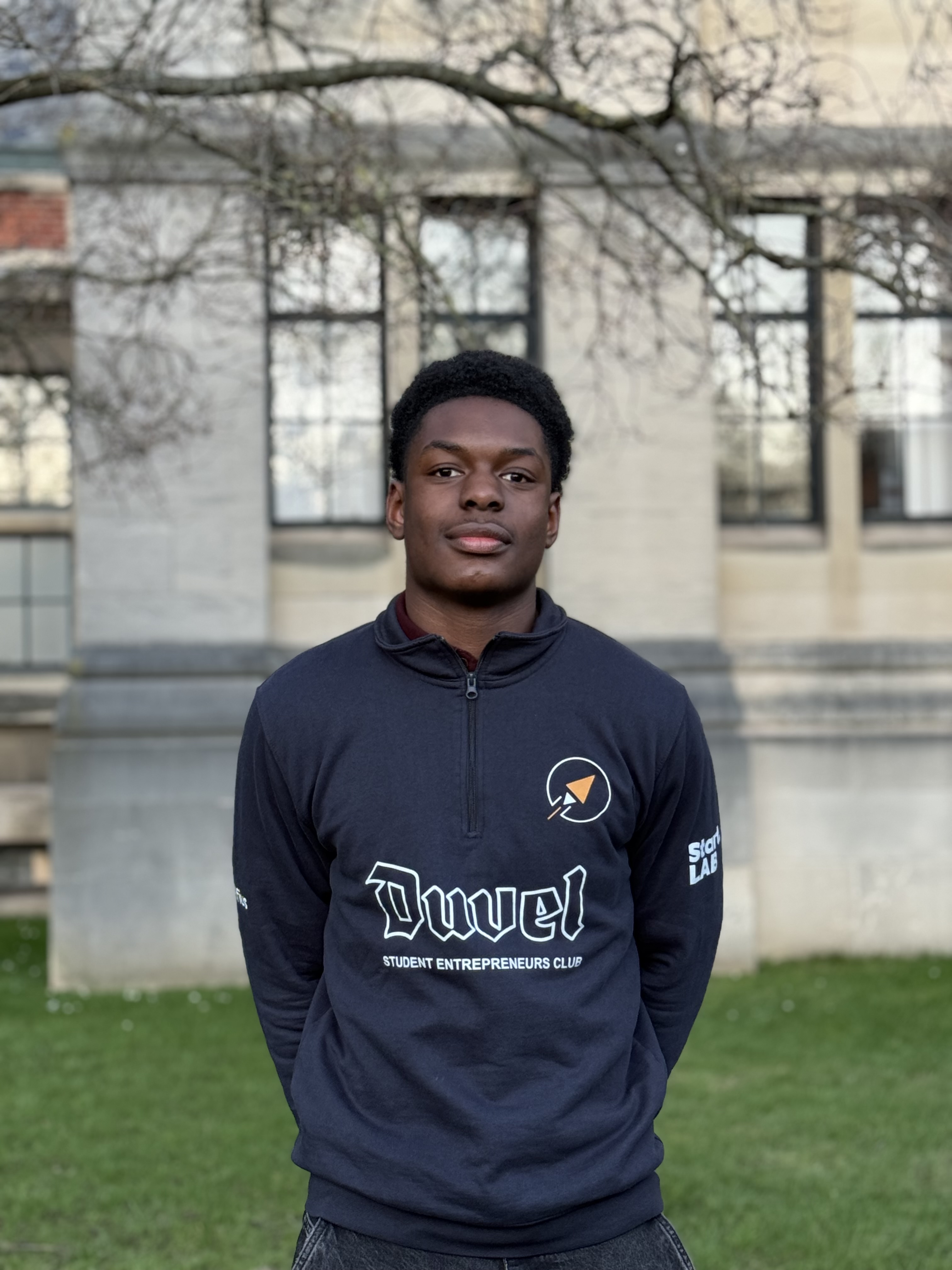 Portrait d'un jeune homme souriant portant un haut noir avec un col montant, sur un fond flou clair.