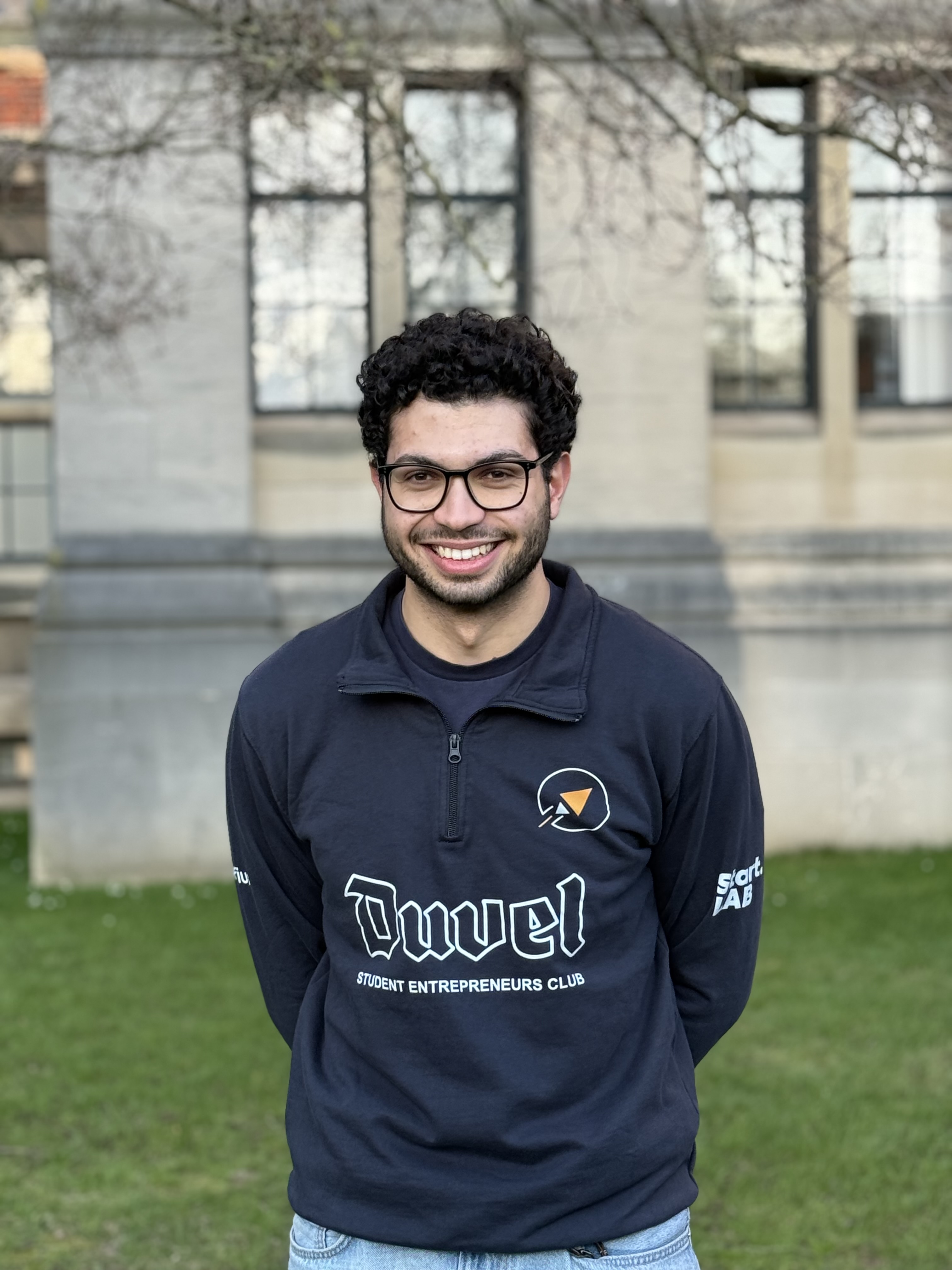 Portrait d'un homme souriant avec des lunettes et des cheveux bouclés foncés, portant un sweat noir et un t-shirt blanc.