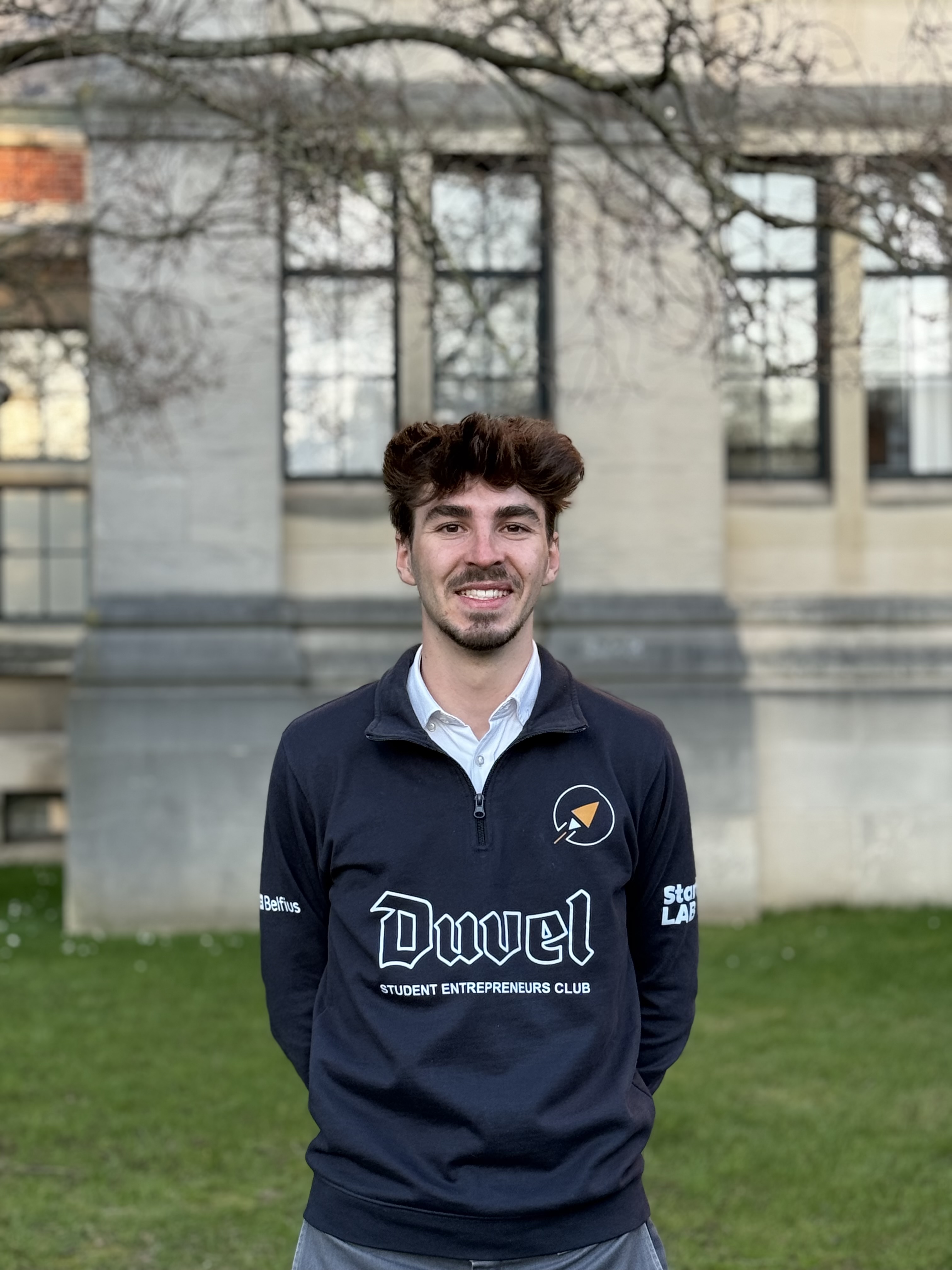 Portrait d'un homme jeune avec une barbe légère et des cheveux bruns ébouriffés, portant un pull noir, sur un fond clair flou.