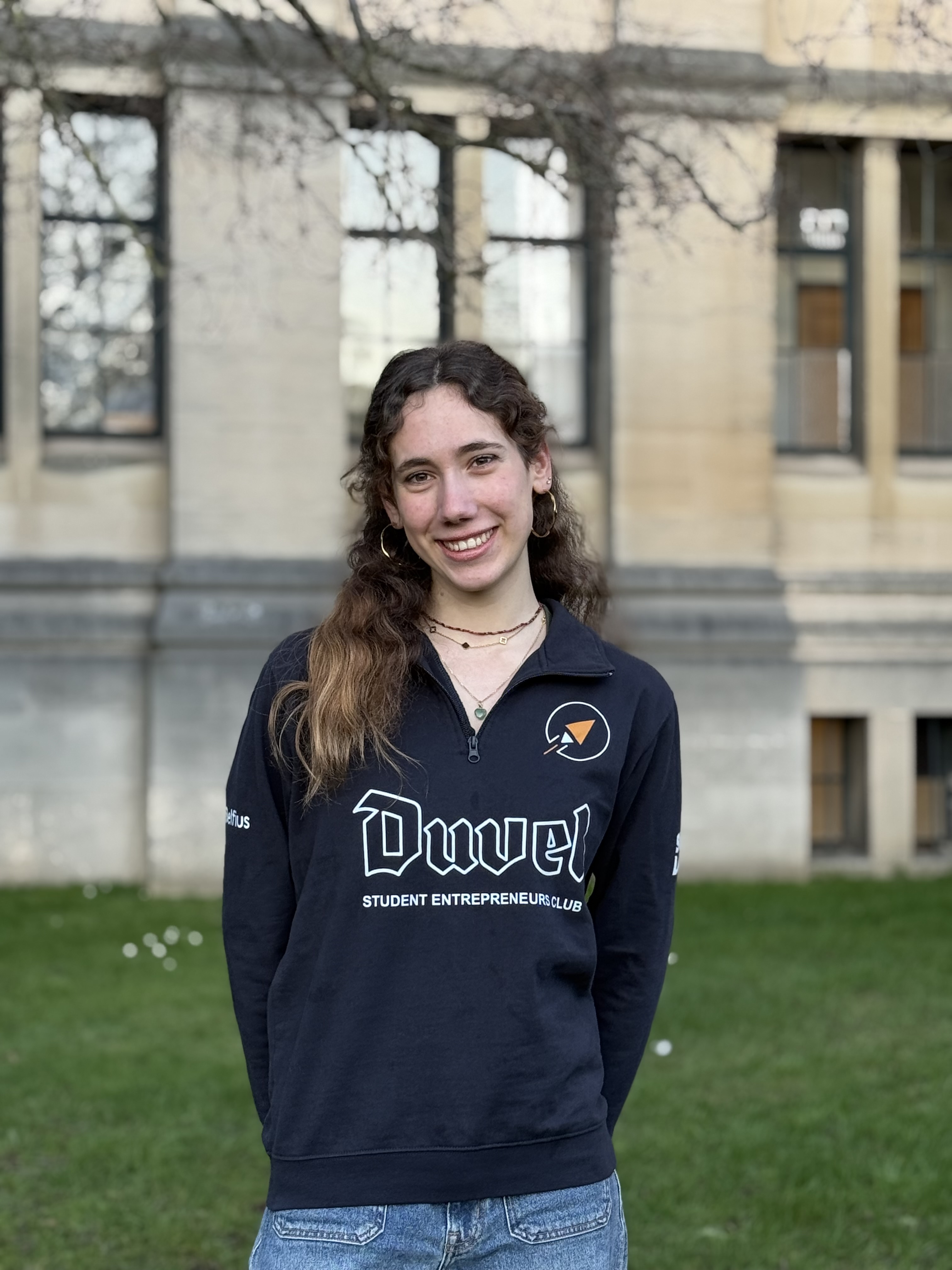 Portrait d'une jeune femme souriante aux cheveux longs et bouclés, portant des boucles d'oreilles et un collier.