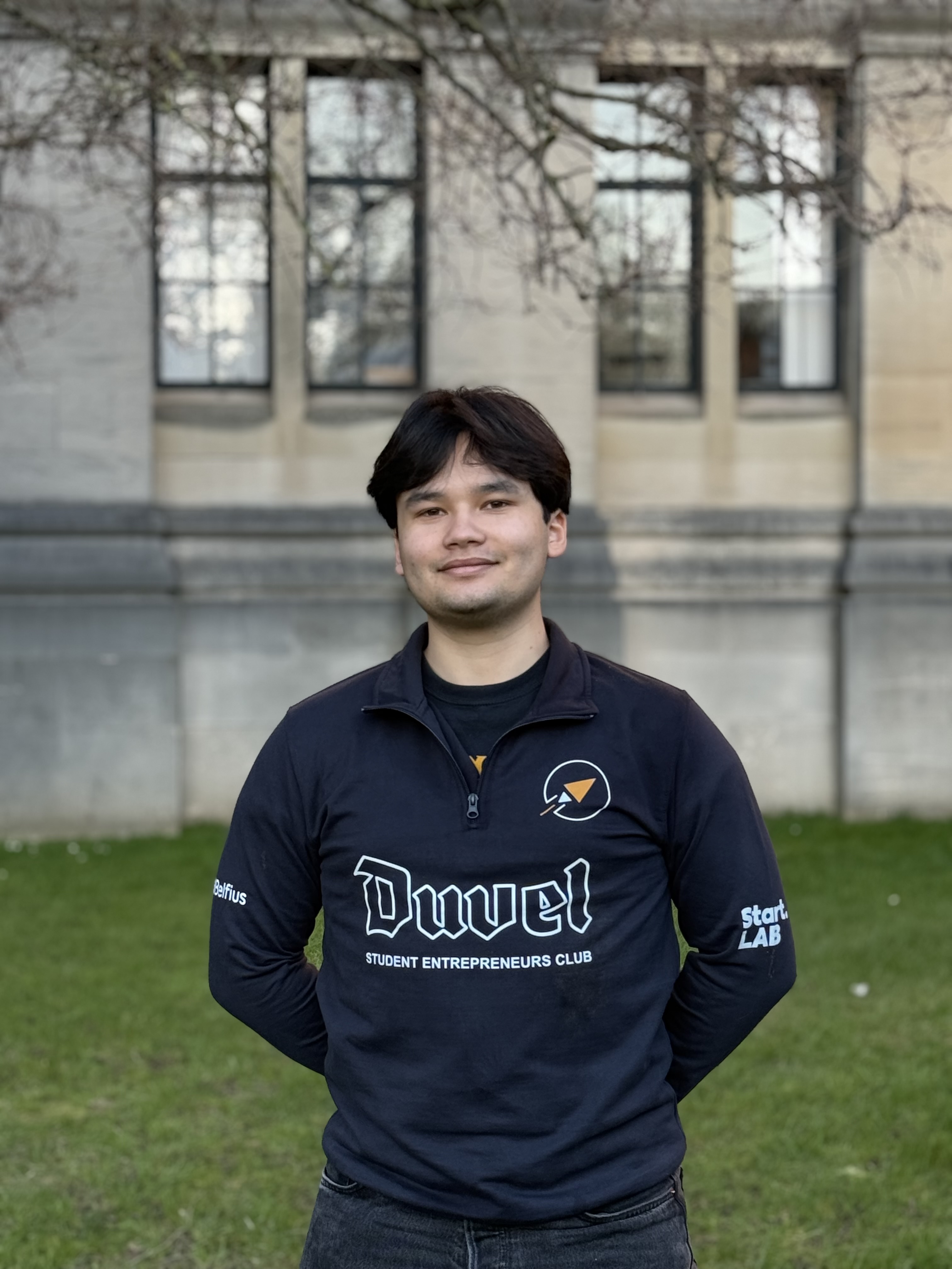 Portrait d'un jeune homme avec des cheveux noirs mi-longs et une expression neutre, portant une veste noire sur un fond clair.