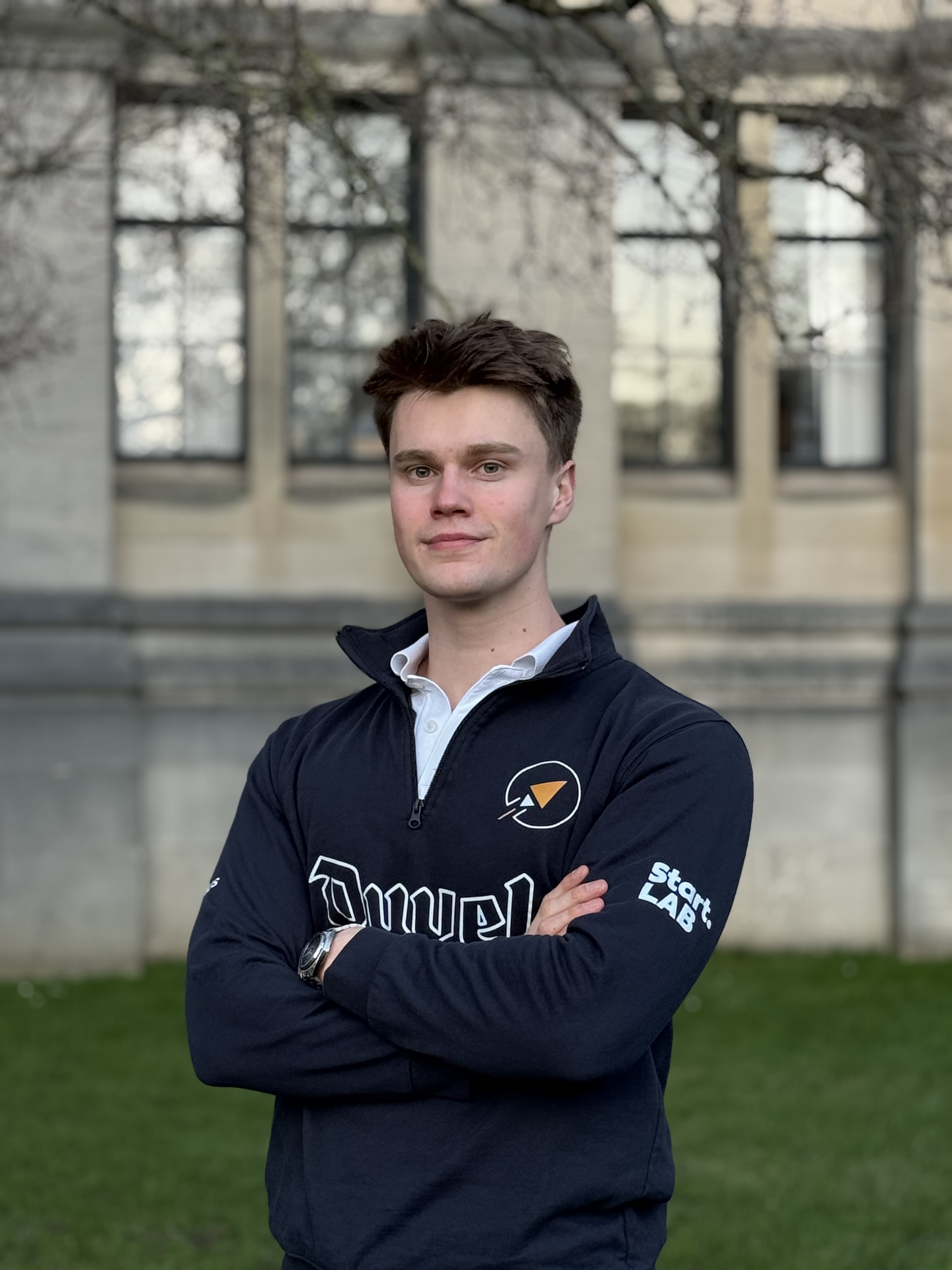 Jeune homme avec un sourire léger, cheveux bruns ébouriffés, éclairage bleu sur un côté du visage, portant un sweat à capuche foncé.