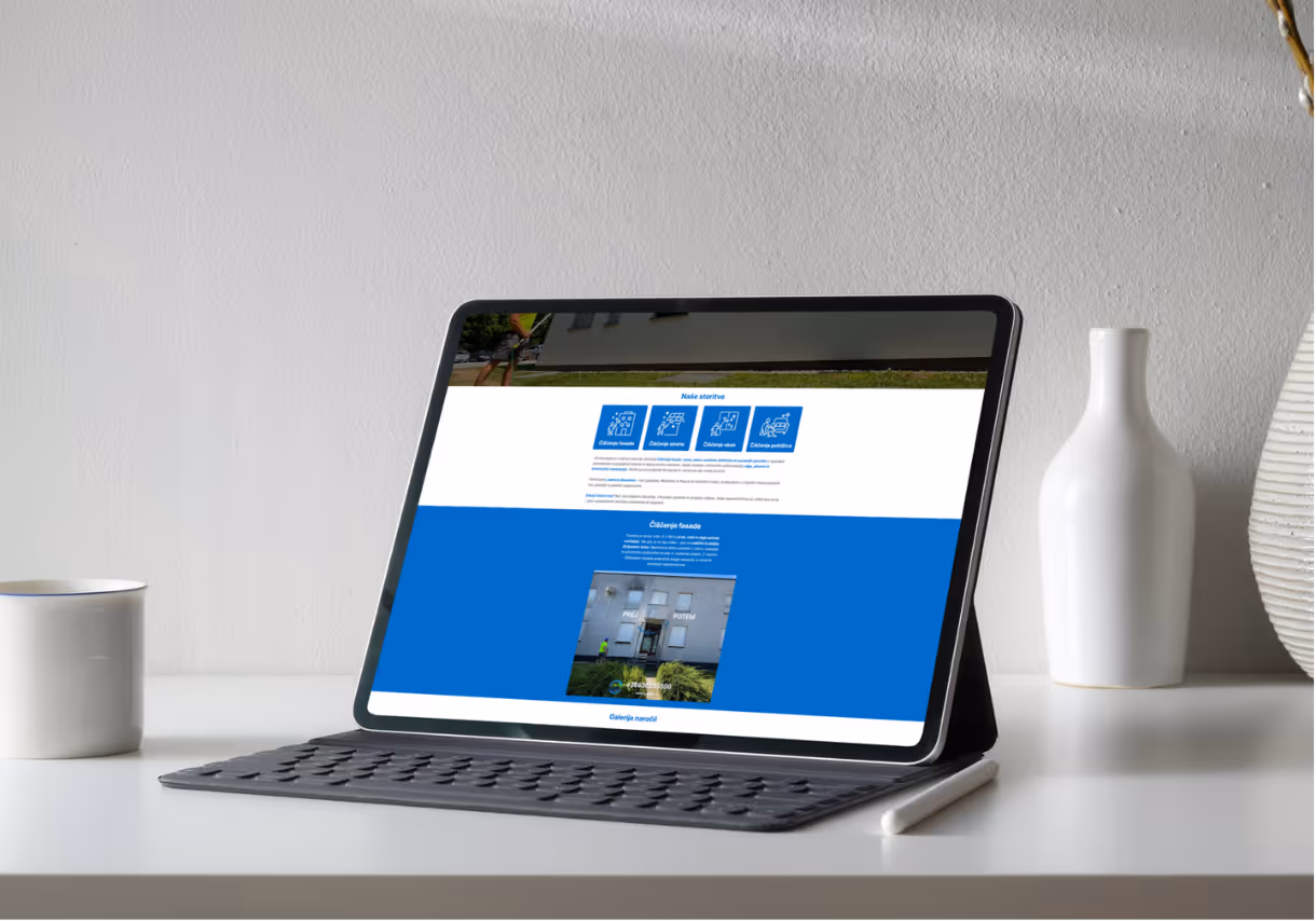 Tablični računalnik z tipkovnico na beli mizi ob beli skodelici in vazi, zaslon prikazuje spletno stran v slovenščini.