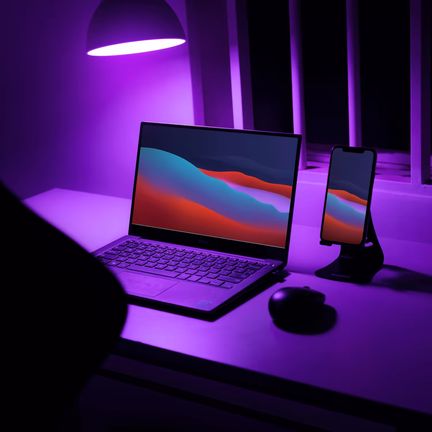 Laptop and smartphone with colorful abstract wallpaper placed on a desk under purple lighting.