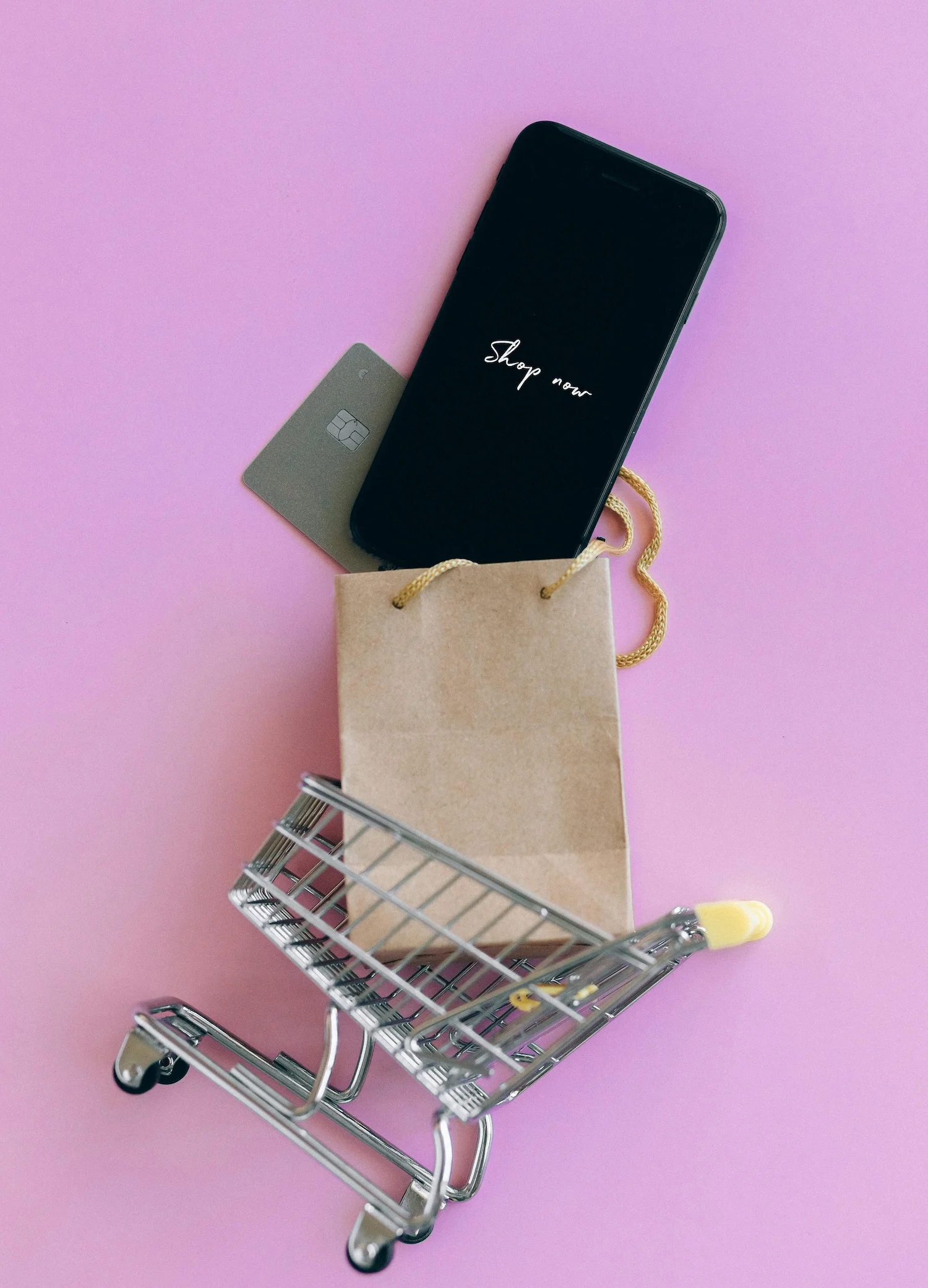 Miniature shopping cart with a brown paper bag, a smartphone displaying 'Shop now,' and a credit card on a pink background.