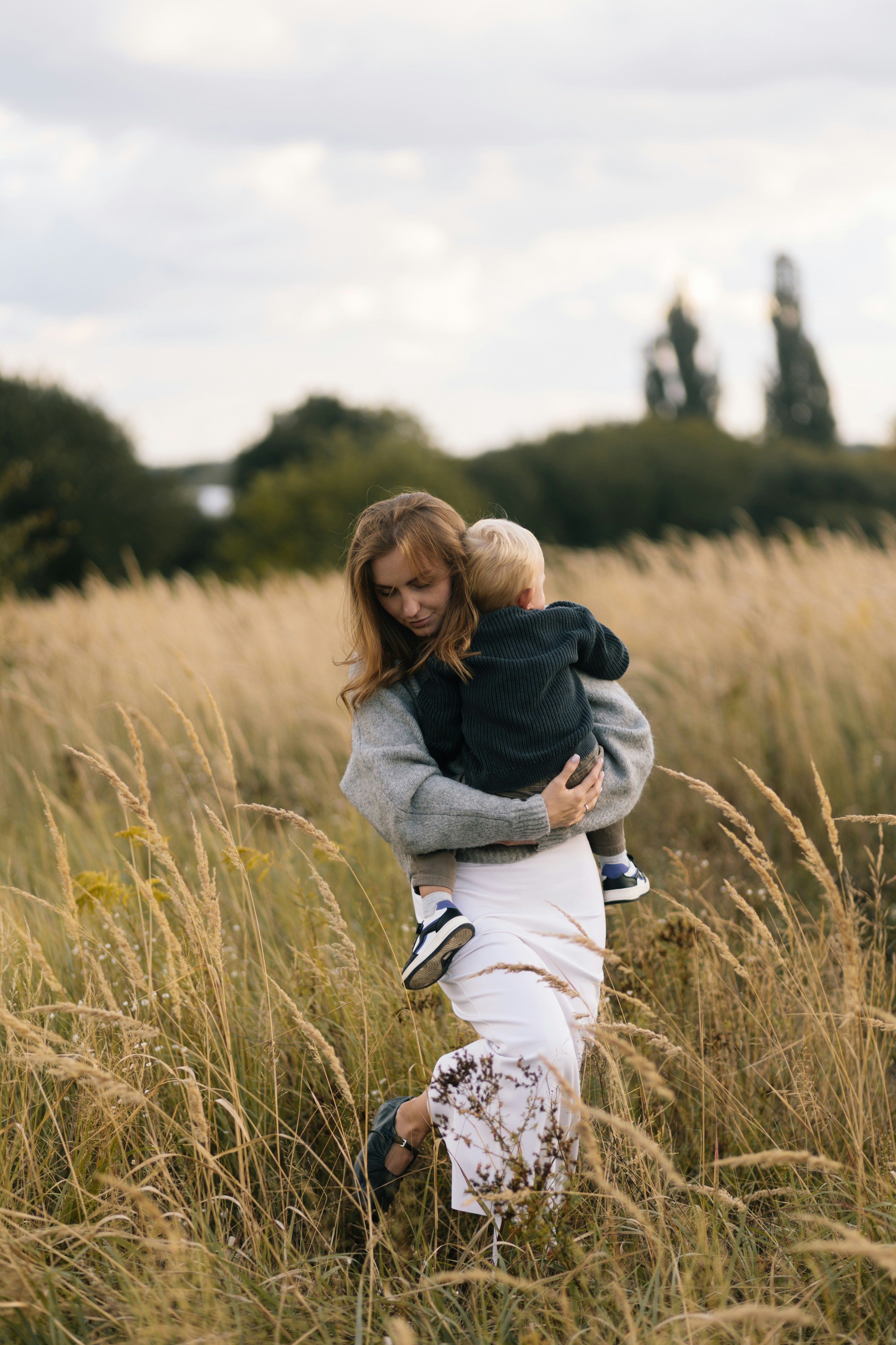 Woman in a gray sweater and white skirt holding and carrying a child in a dark sweater through a tall grassy field.