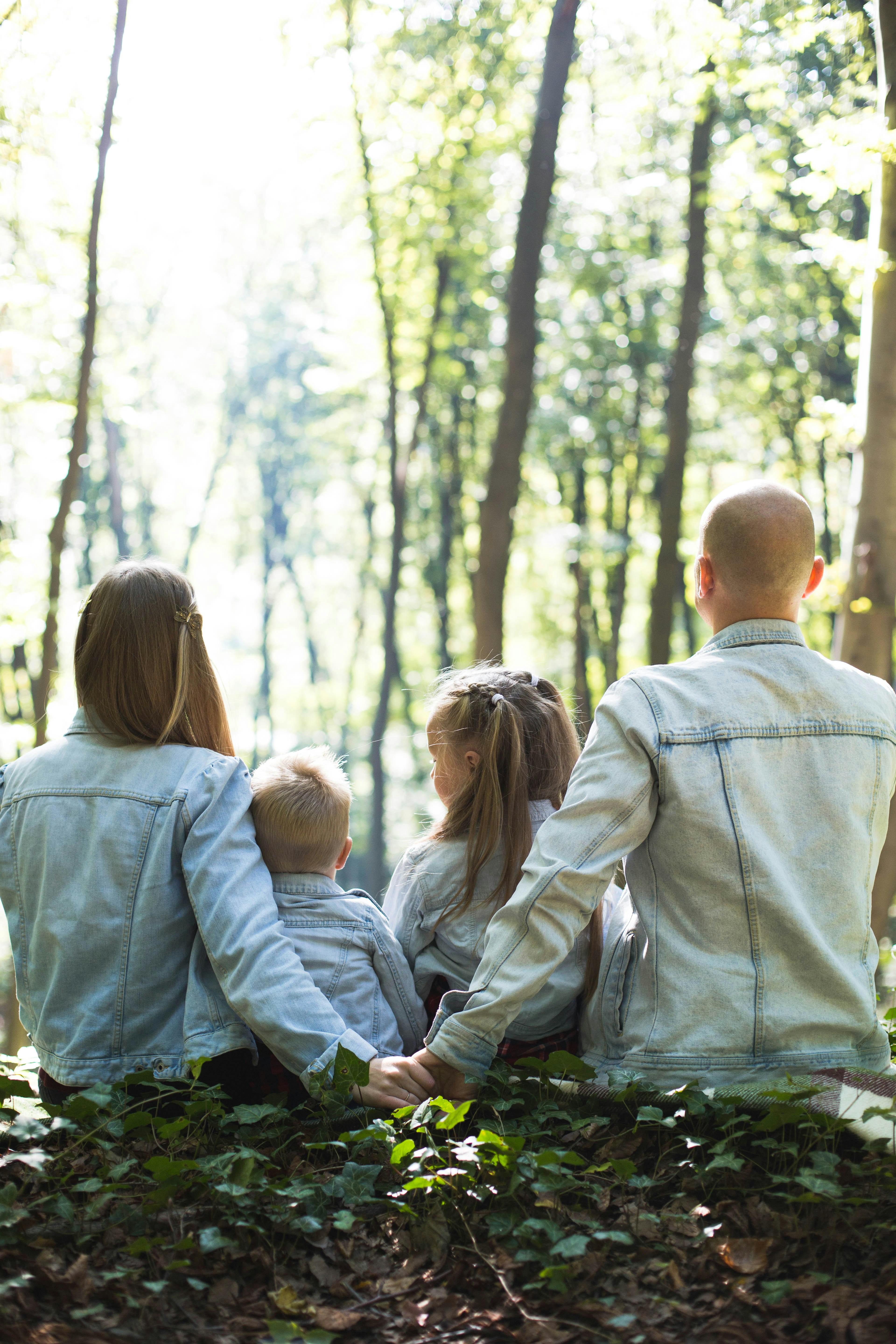 Family of four sitting on the forest ground holding hands and wearing denim jackets, facing away.