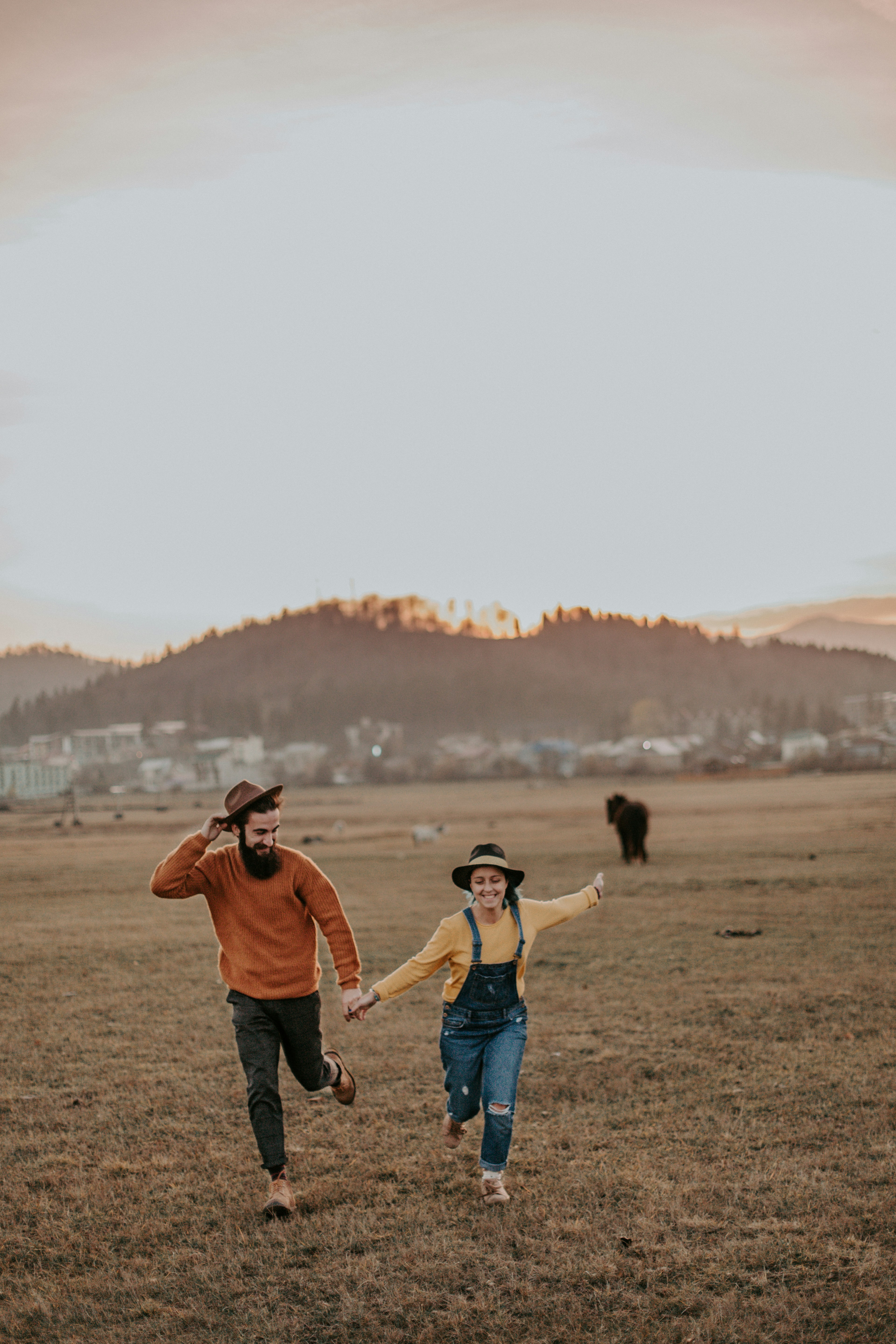 Man and woman holding hands and running in a field at sunset, with hills and distant buildings in the background.