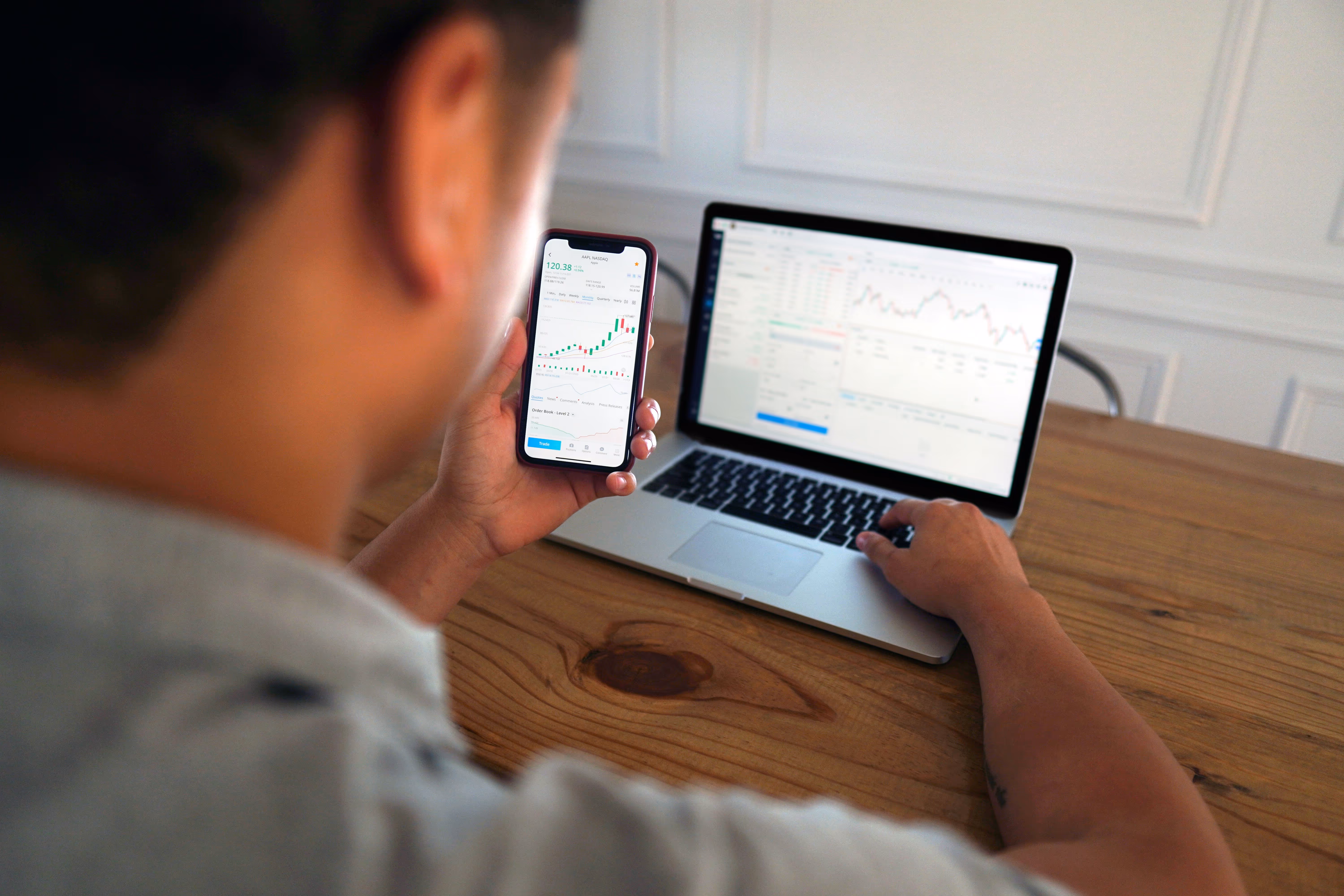 Person using a smartphone showing a stock market chart while working on a laptop with financial data displayed.