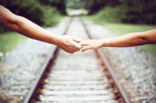 Two people holding hands over railroad tracks, with engagement ring visible on one hand.