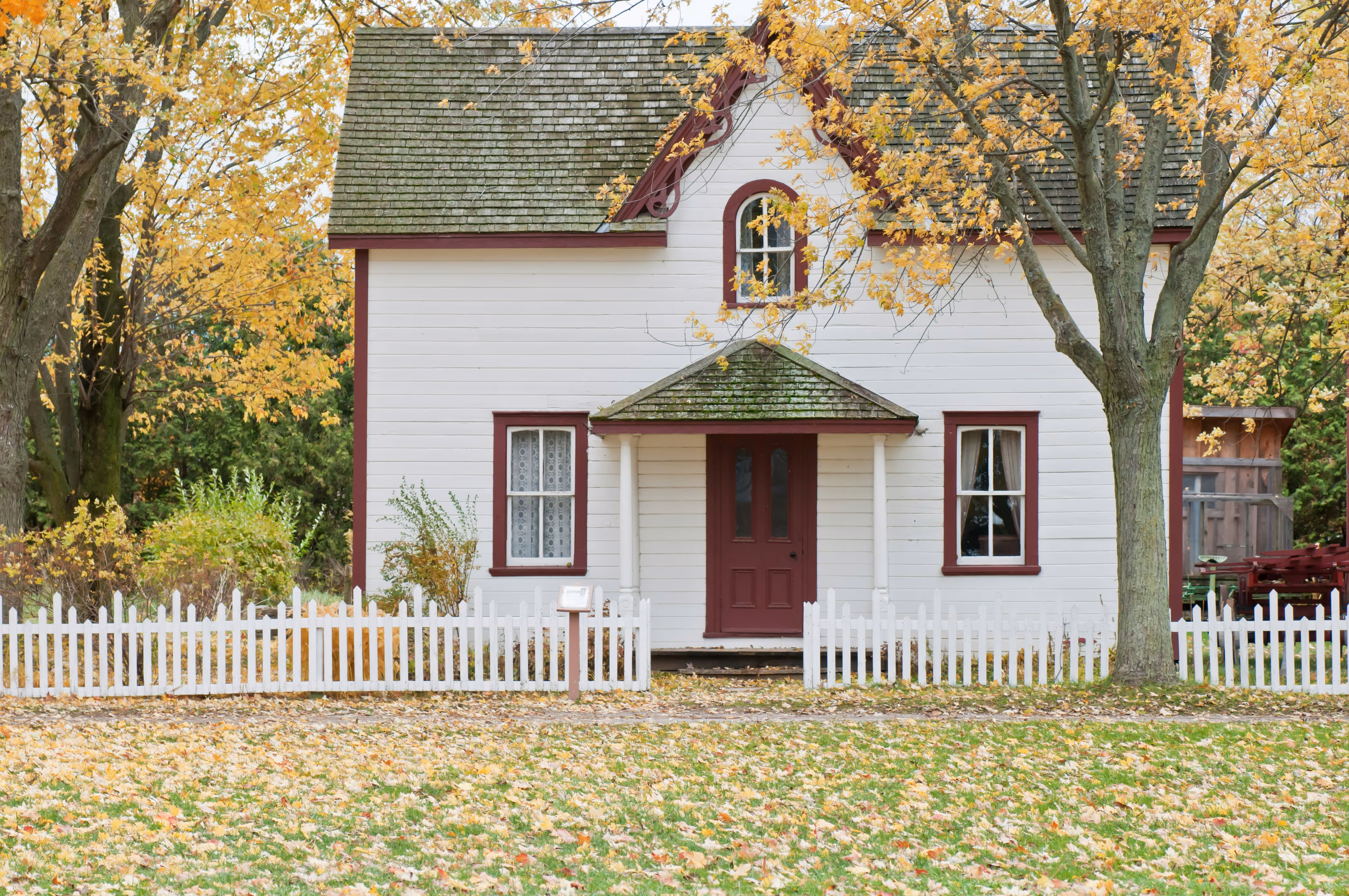 White cottage with maroon trim surrounded by autumn trees and a white picket fence.