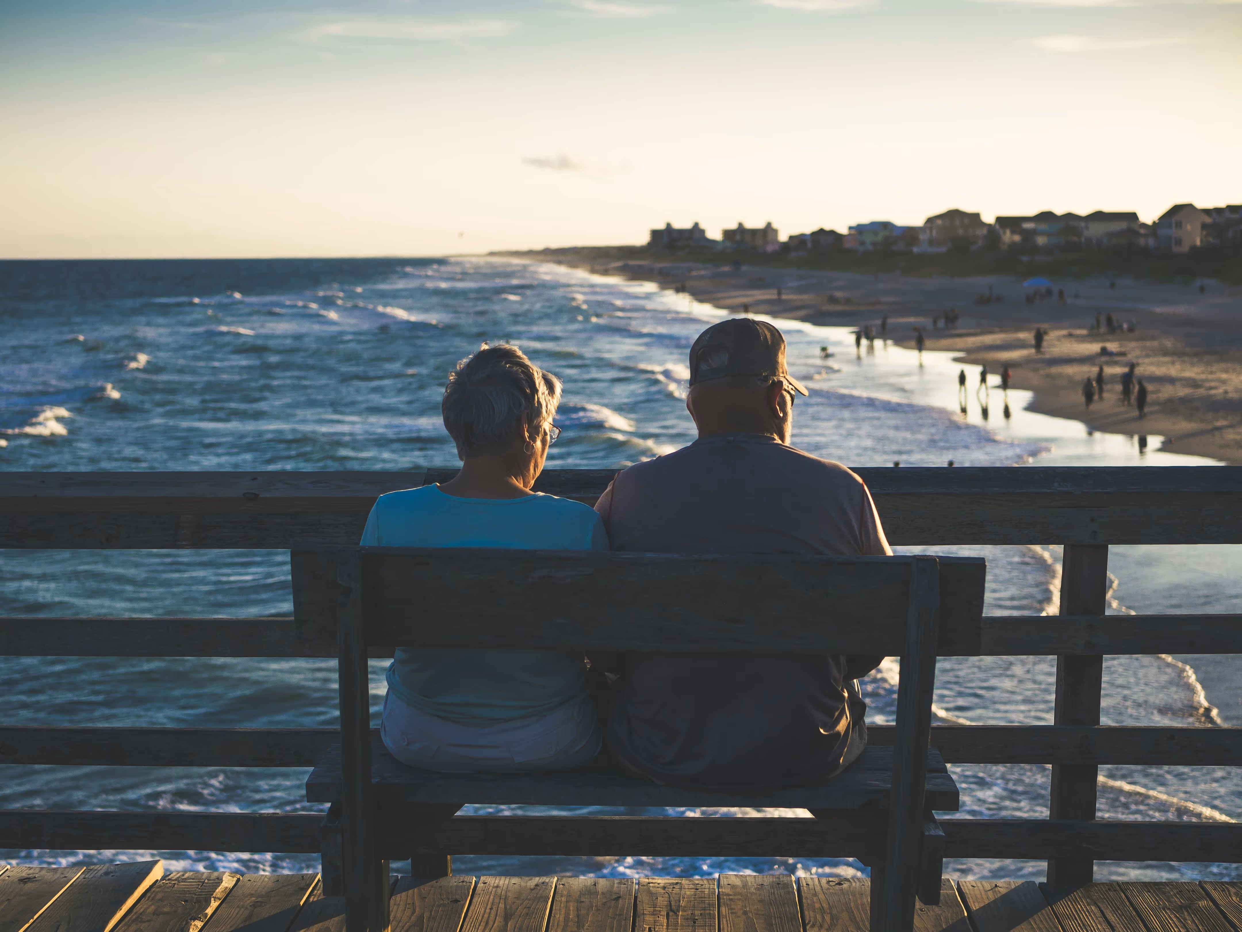 An elderly couple sitting on a wooden bench on a pier, overlooking the ocean and a beach with people in the distance during sunset.