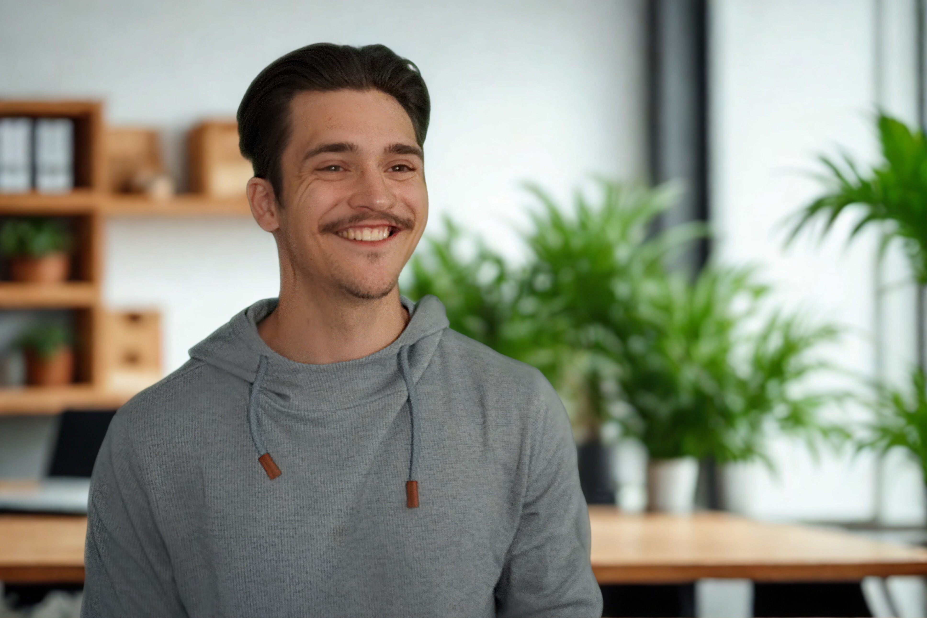 Smiling young man with dark hair and a mustache wearing a gray hoodie in a modern office with plants and wooden shelves.