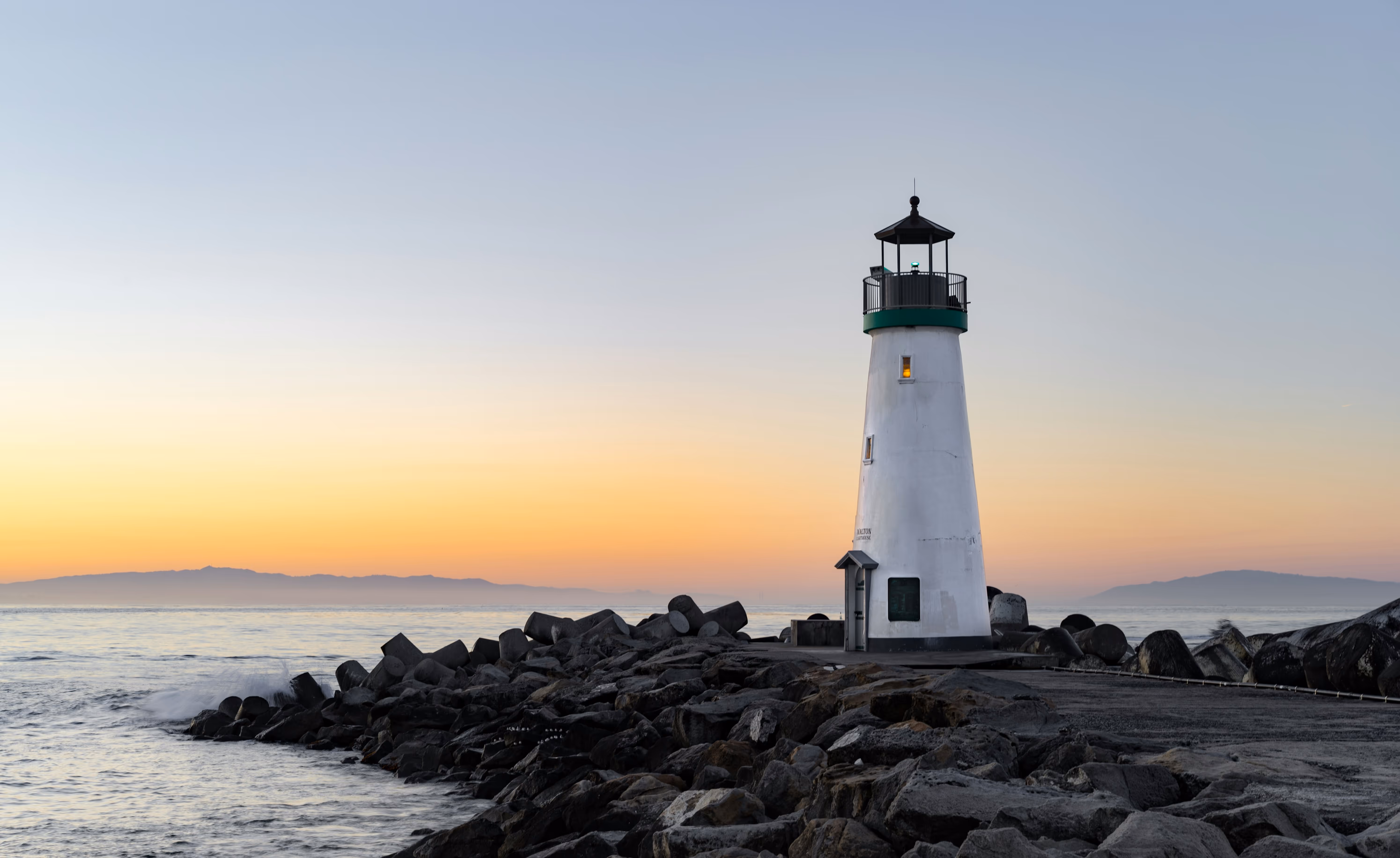 White lighthouse on rocky pier with calm ocean and sunset sky in the background.