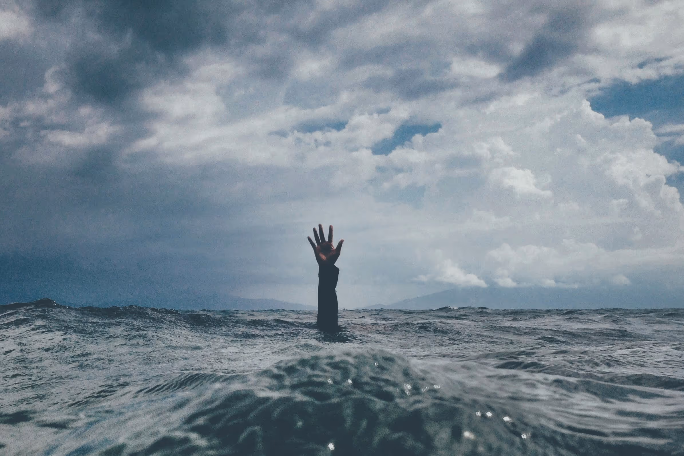 A single hand reaching out of rough ocean waves under a cloudy sky.