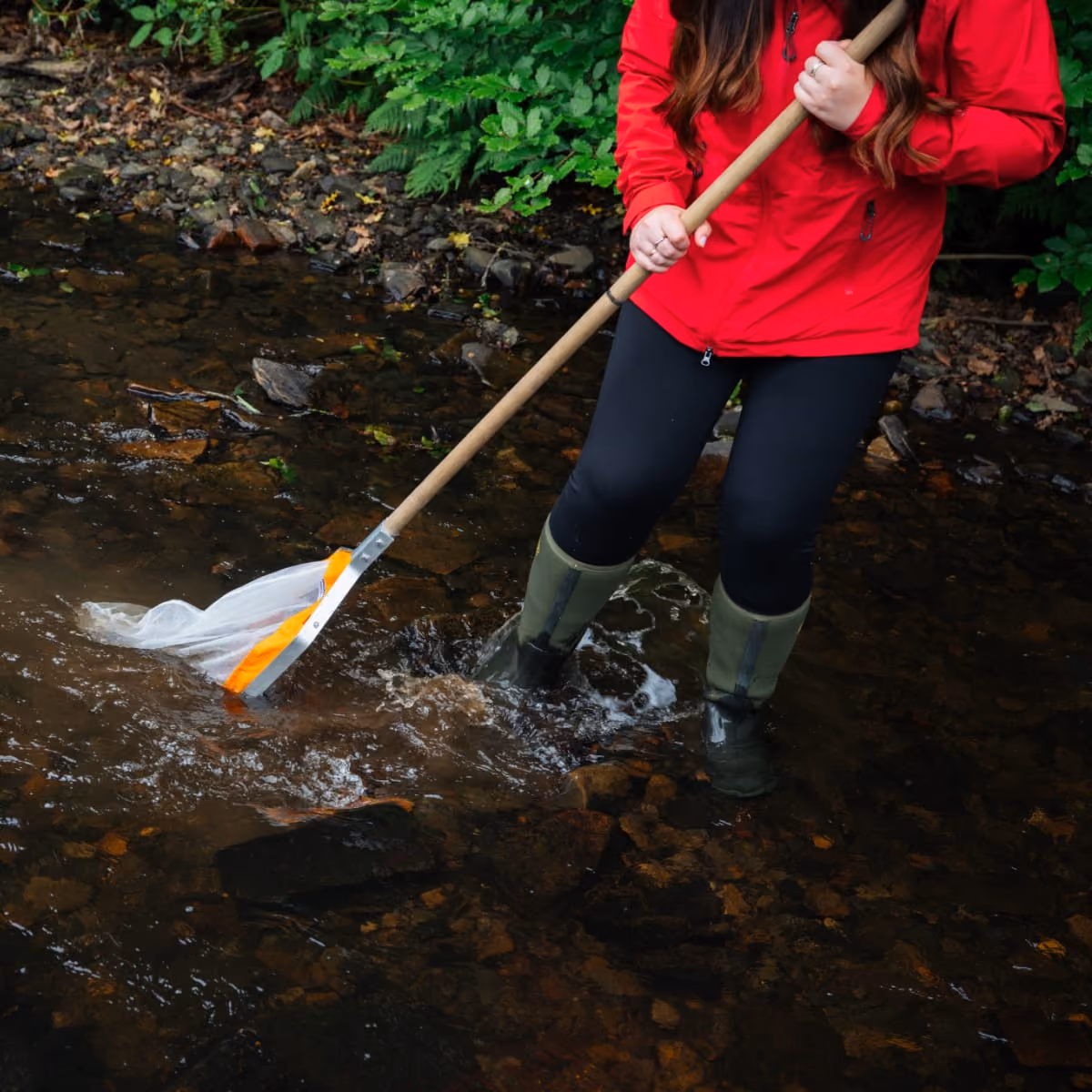 Person in red jacket and green boots standing in water using a long-handled net to collect samples.