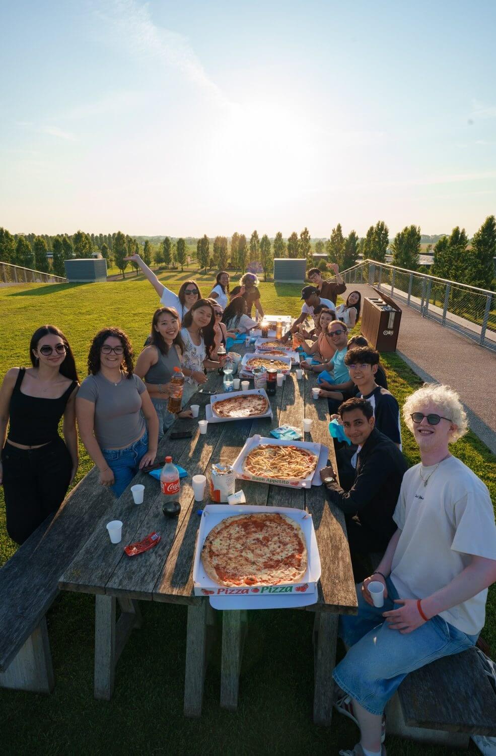 Group of young people enjoying an outdoor picnic around a long wooden table with pizzas and drinks at sunset.