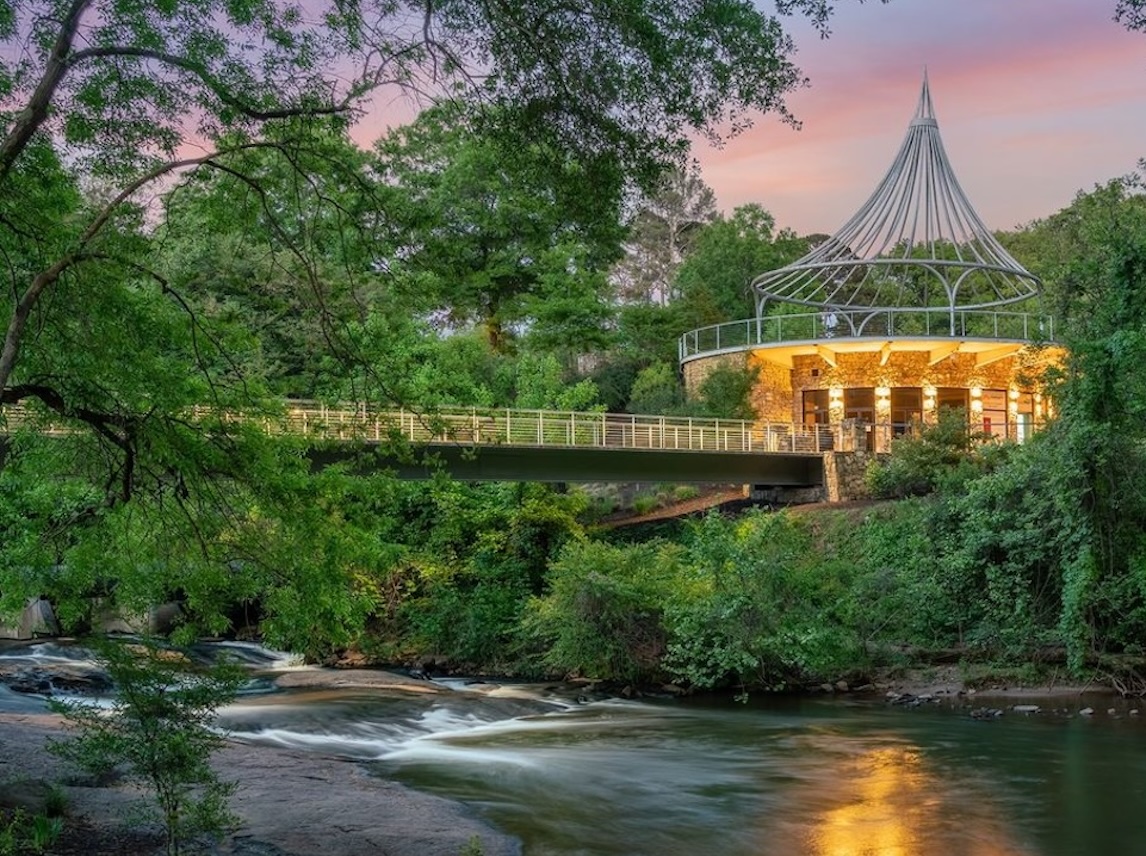 Stone building with illuminated windows and metal canopy next to a bridge over a flowing river surrounded by dense green trees at sunset.