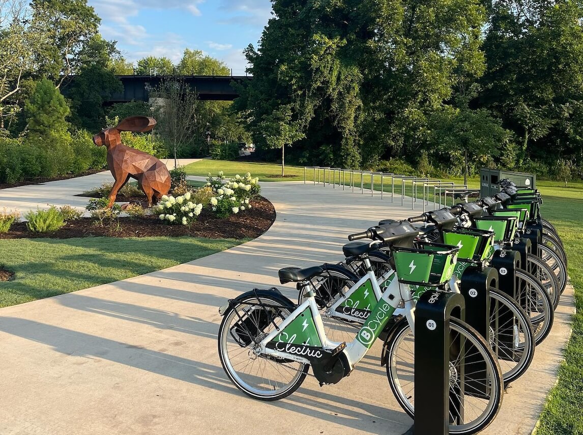 Row of green and white electric rental bicycles parked on a concrete path near a park with a large metal rabbit sculpture and trees.