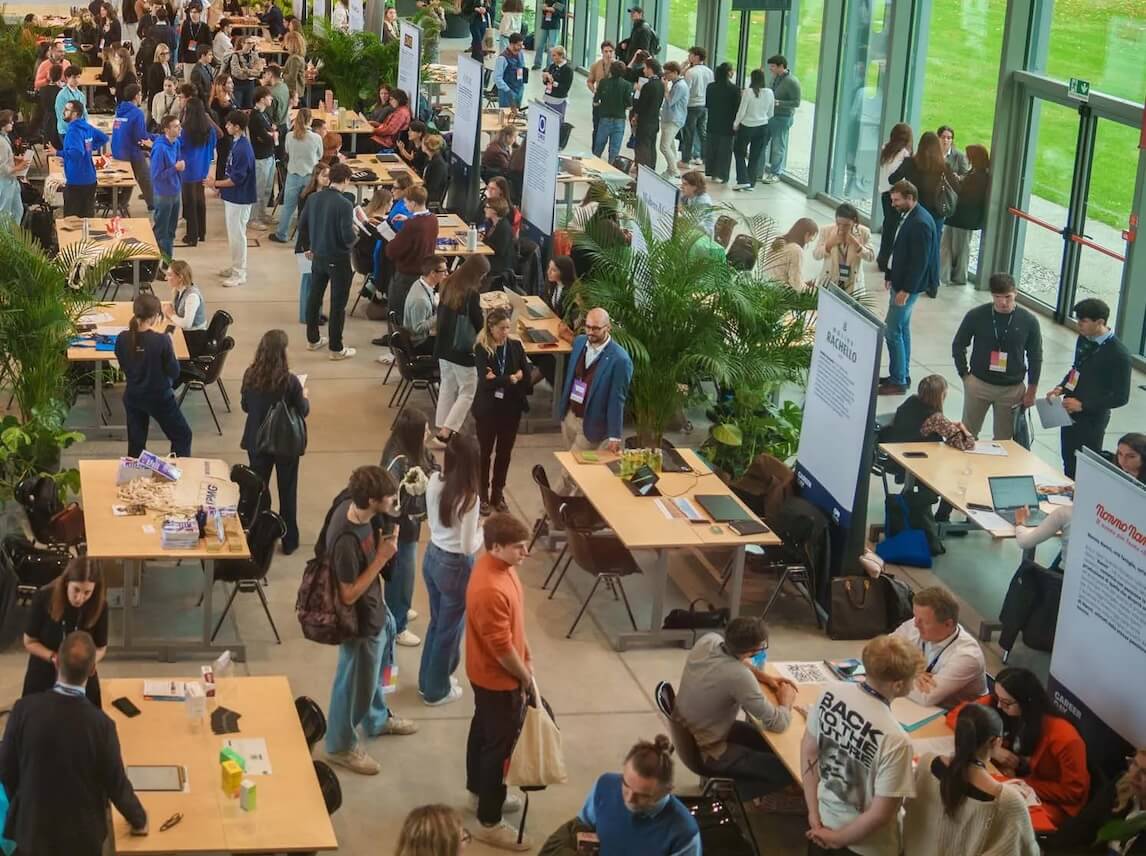 Crowded indoor event with people networking and sitting at tables with laptops and promotional materials in a bright room with large windows.