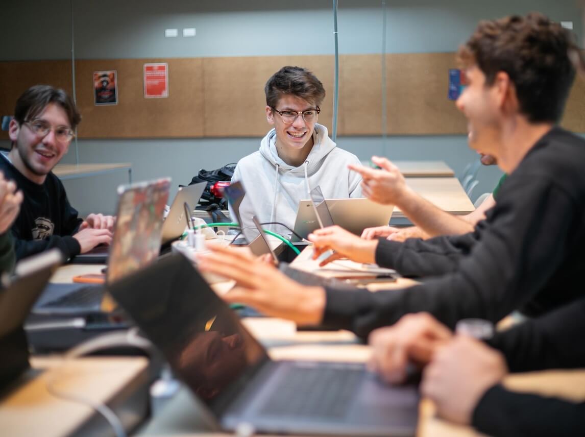 A group of young people sitting around a table with laptops, smiling and talking in a casual meeting or study session.