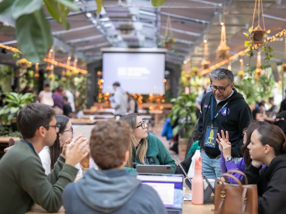 Group of people engaged in a discussion around a table in a green-themed, well-lit indoor space with hanging plants and a projection screen in the background.
