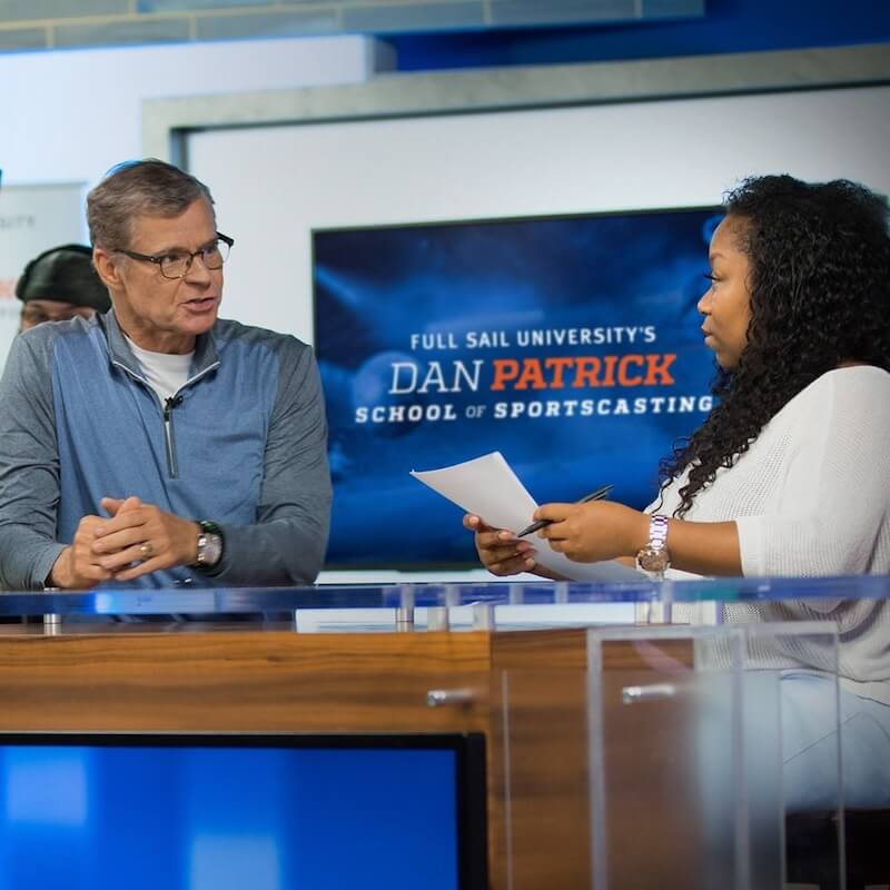 A man and woman engaged in a discussion on a set with a screen behind them reading 'Full Sail University's Dan Patrick School of Sportscasting'.