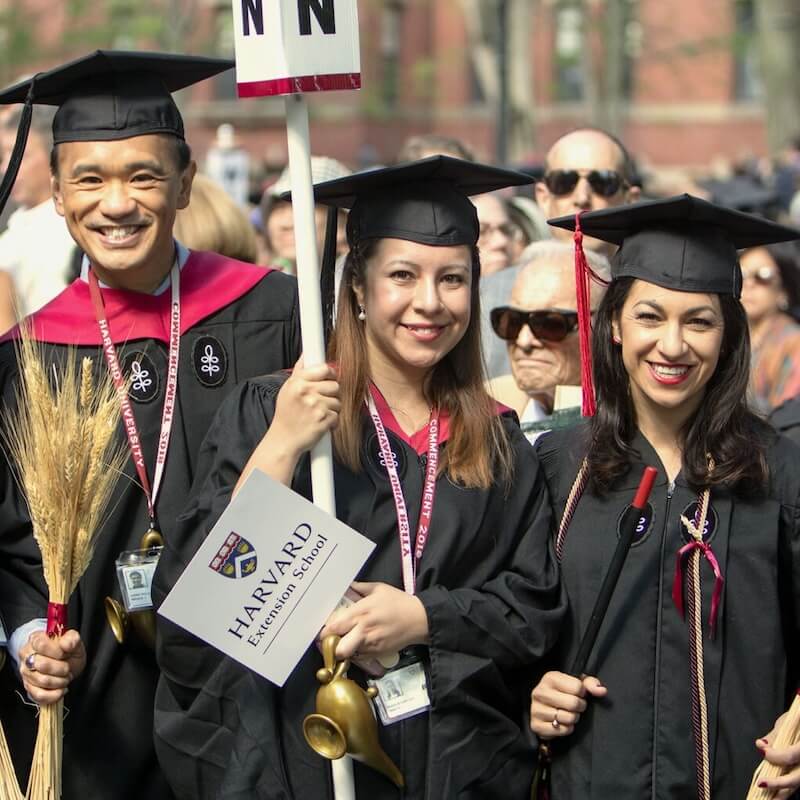 Three Harvard Extension School graduates in caps and gowns smiling during a commencement ceremony, holding celebratory items.