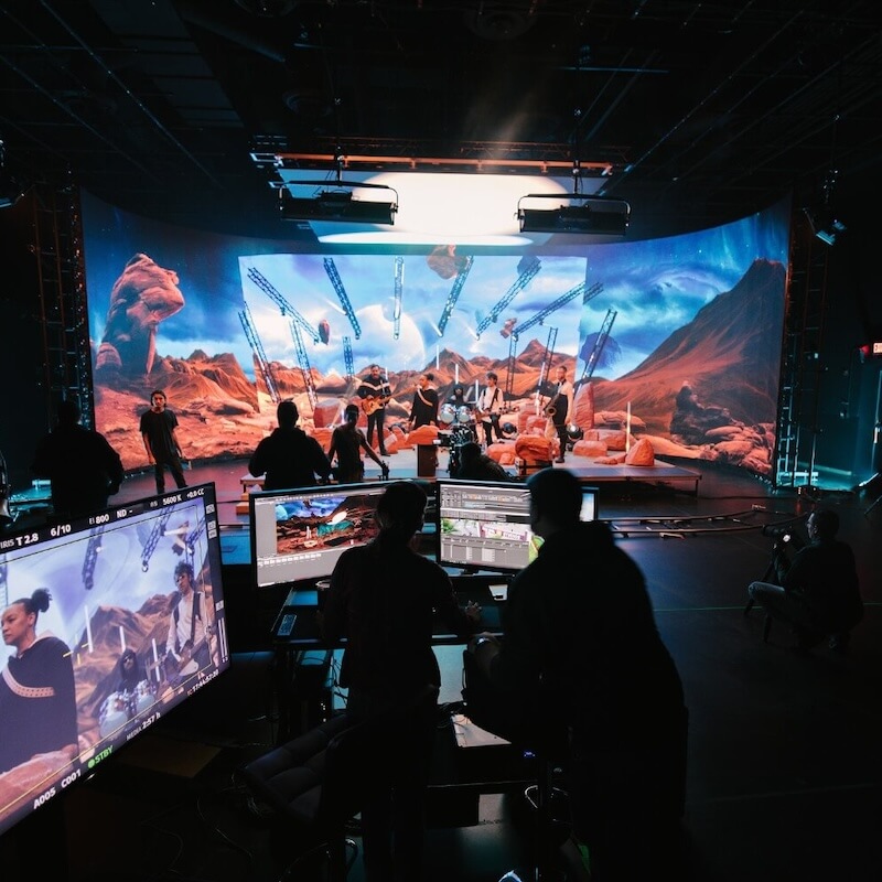 Film crew filming musicians on a stage with a large desert and mountain virtual background on screens.