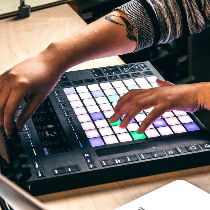 Person using a music production controller with lit-up multi-colored pads and knobs on a wooden surface.