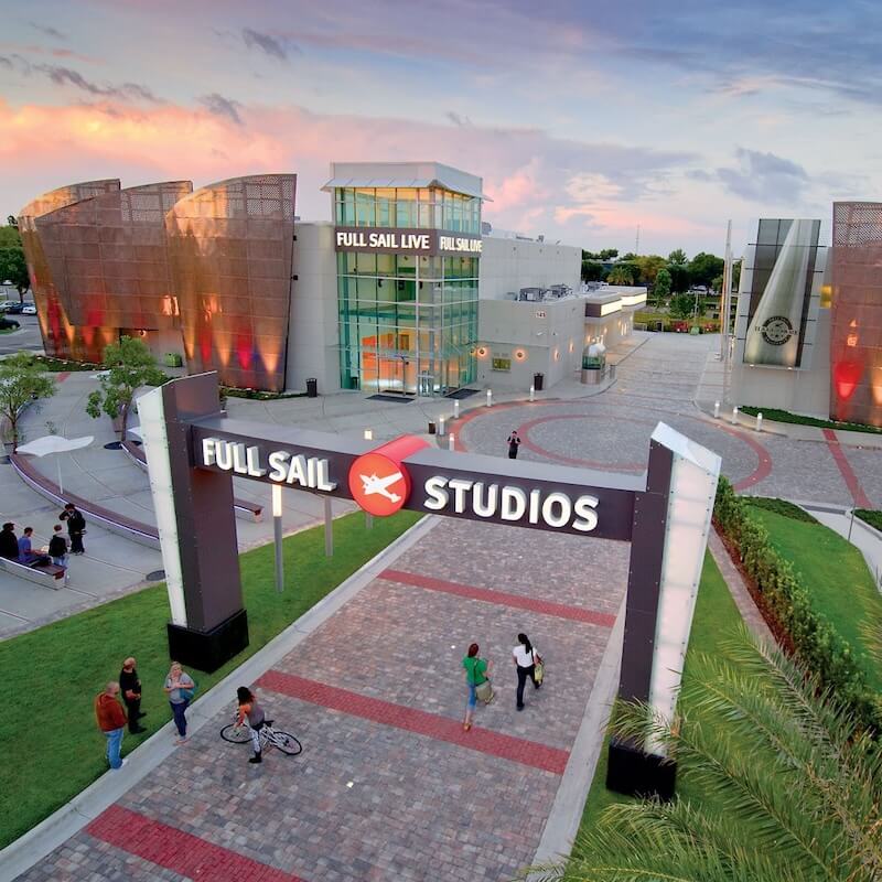 Entrance archway reading 'Full Sail Studios' over a paved pathway leading to a modern building with glass and copper-colored architectural elements during sunset.