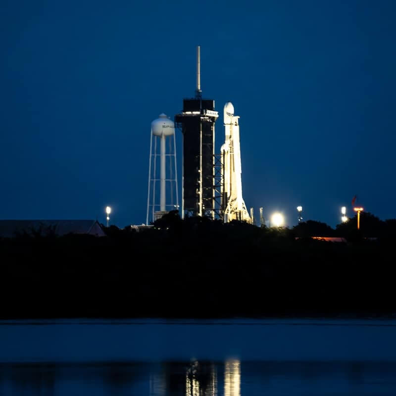 Illuminated space rocket on launch pad at night with reflection on water.
