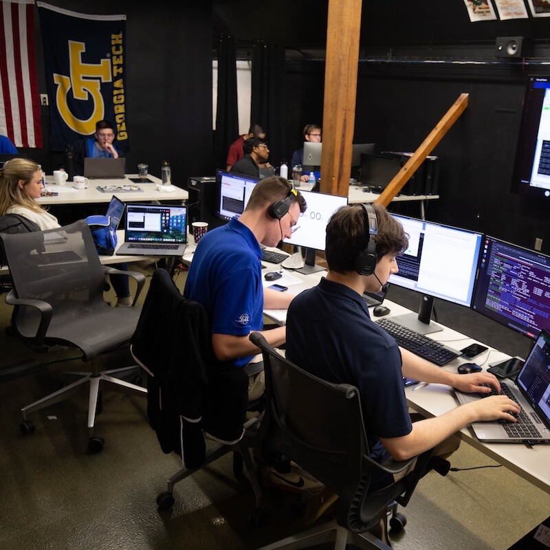 Group of people working on laptops and desktop computers in an office with Georgia Tech flag and American flag on the wall.