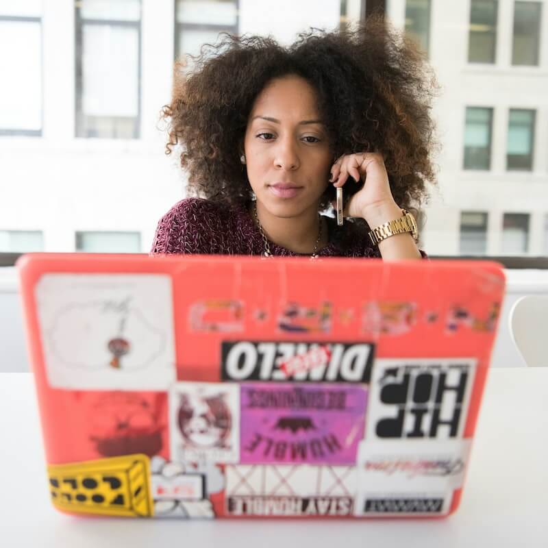 Woman with curly hair talking on a smartphone while working on a laptop covered with various colorful stickers.
