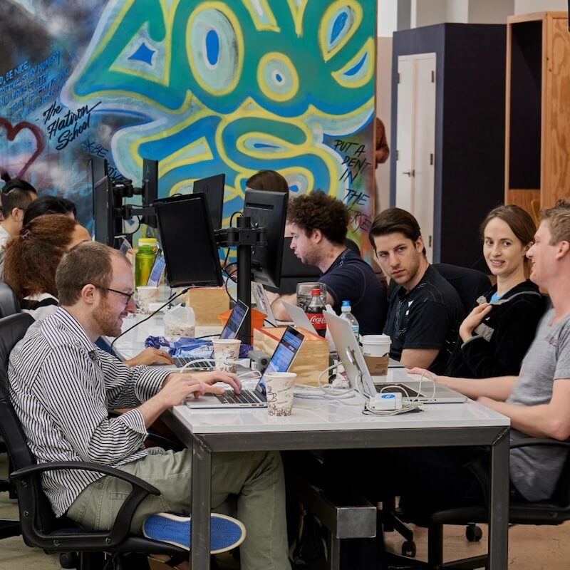 Group of people working on laptops and desktop computers around a shared table in a modern office with colorful graffiti on the wall.