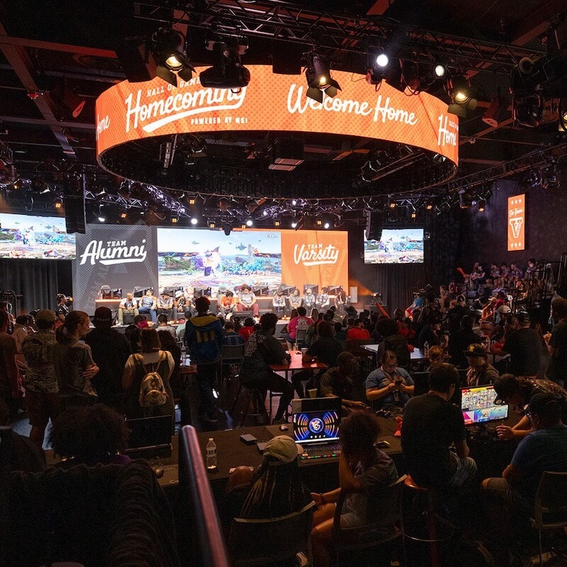 Large indoor esports event with teams labeled Alumni and Varsity competing on stage in front of a seated audience.